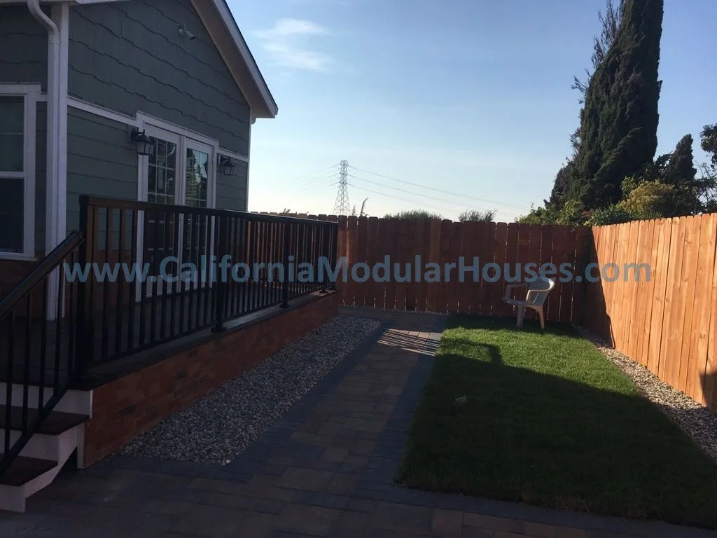 Backyard with a wooden fence, a small grassy patch, a paved walkway, and a white plastic chair, adjacent to a house with gray siding and a deck with black railing.