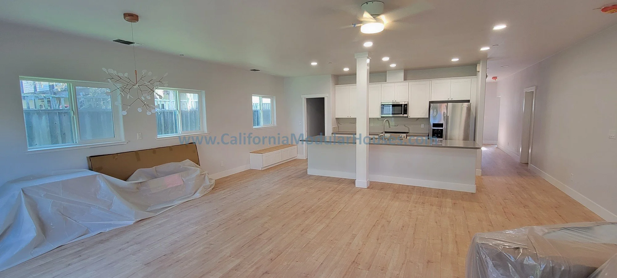 Empty, newly renovated open-concept living space with white walls, light wood flooring, and large windows. Kitchen features white cabinets, stainless steel appliances, and a small peninsula. There is a modern chandelier in the living area. Plastic co