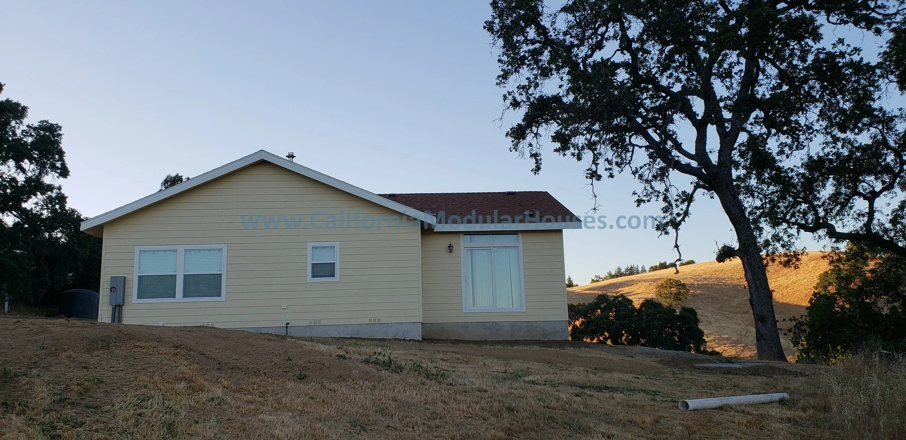 A yellow house with white trim and a brown roof sits on a dry, grassy hill with a large tree nearby, under a clear sky at sunset. Bay Area Modular Home, San Jose Modular Homes, Modular Home California,