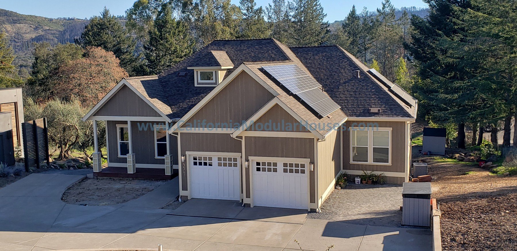 A two-story modern house with solar panels on the roof surrounded by trees and hills in the background. The house has a front porch, a double garage, and a gravel driveway.  Santa Rosa, CA.  Sonoma County.  California Modular, California Modular Home