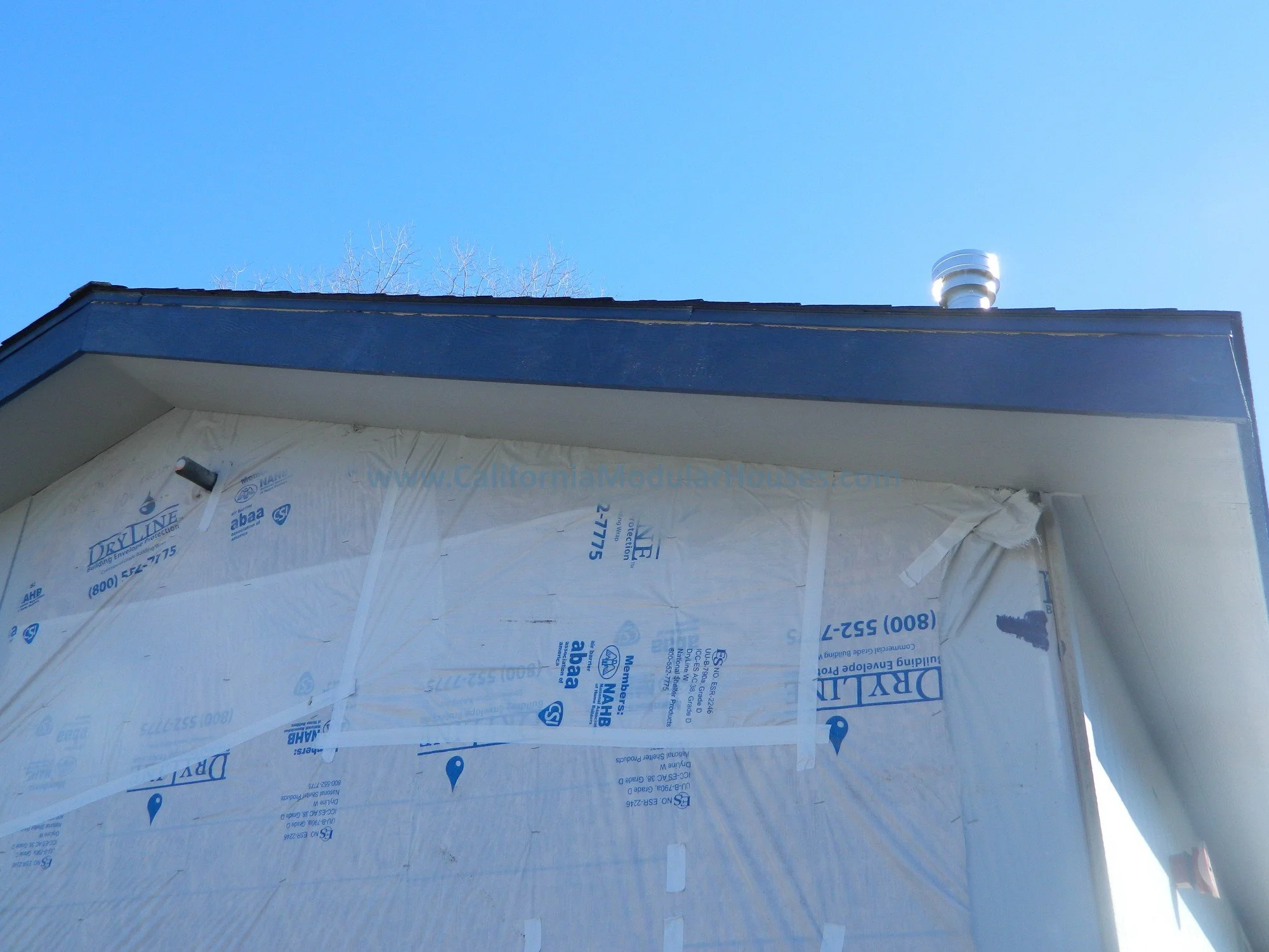 Close-up of a house under construction with white weather-resistant barrier and black roof, against a clear blue sky, with building vents visible on the roof.