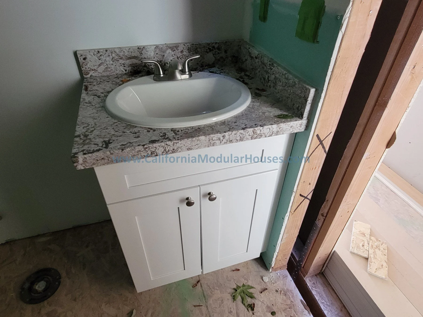 Bathroom vanity with a white cabinet, a granite countertop, and an oval white sink with a silver faucet. Partially framed wall and construction materials are visible.