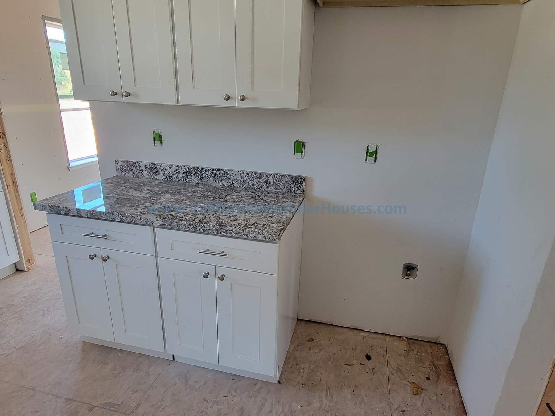Kitchen area under renovation with white cabinets, granite countertop, and empty wall with electrical outlets.
