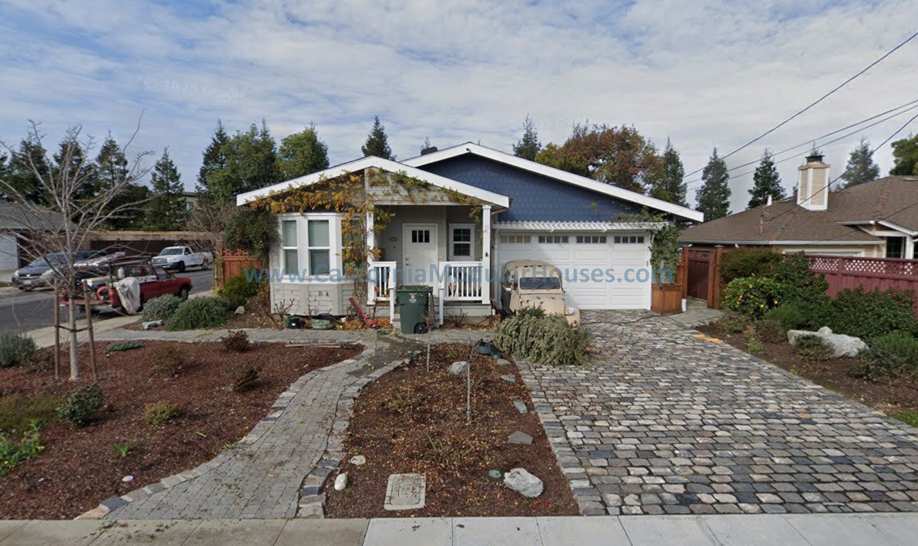 Front view of a small house with a white garage door, blue siding, white trim, a small front porch, and a cobblestone driveway. There are plants, trees, and shrubs in the yard.  San Mateo City, San Mateo County, CA.   California Modular Homes.