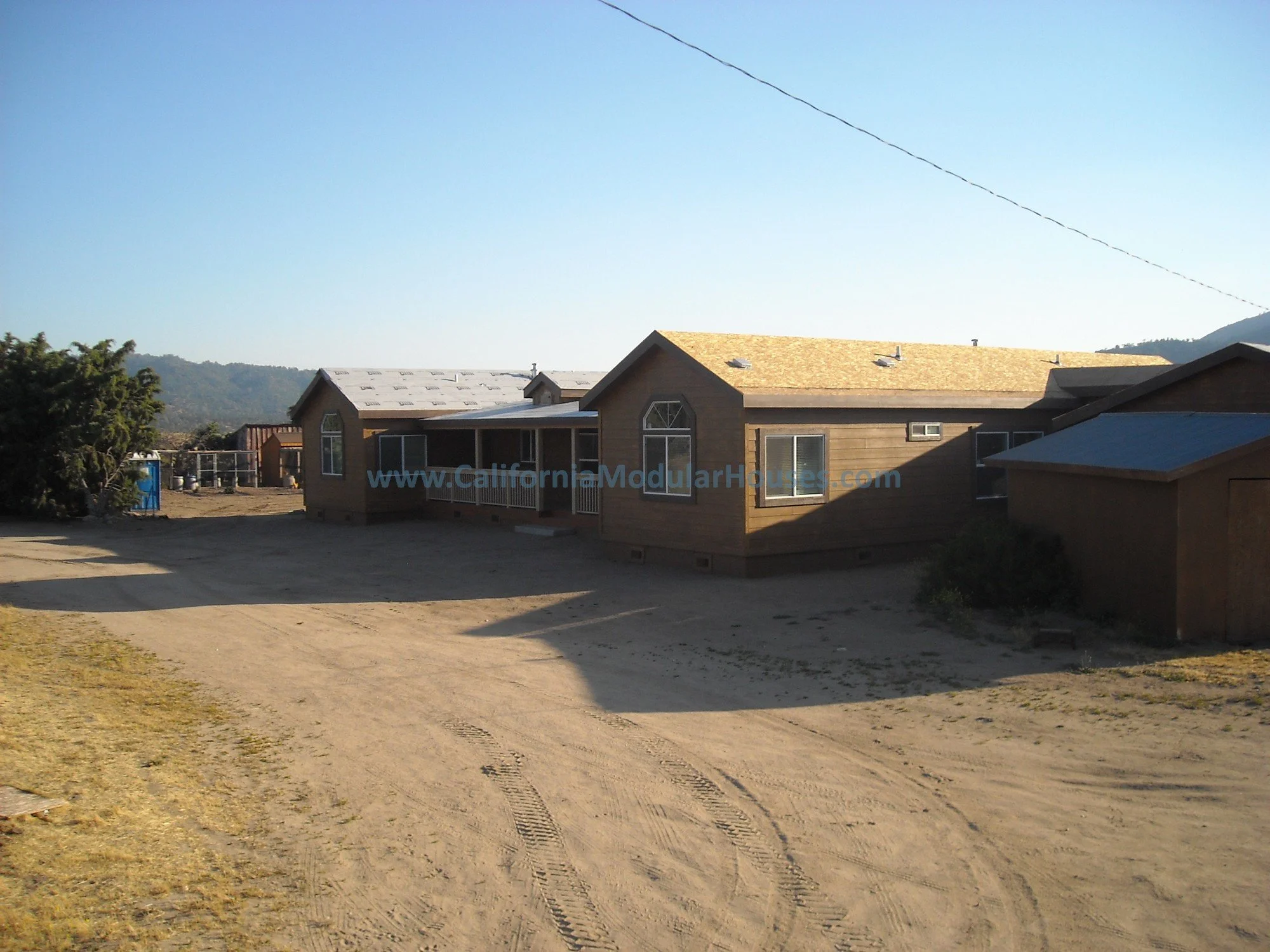 A single-story house with a brown exterior, multiple windows, and a porch, situated on a dirt lot with tire tracks. Mountainous landscape in the background and a blue sky.