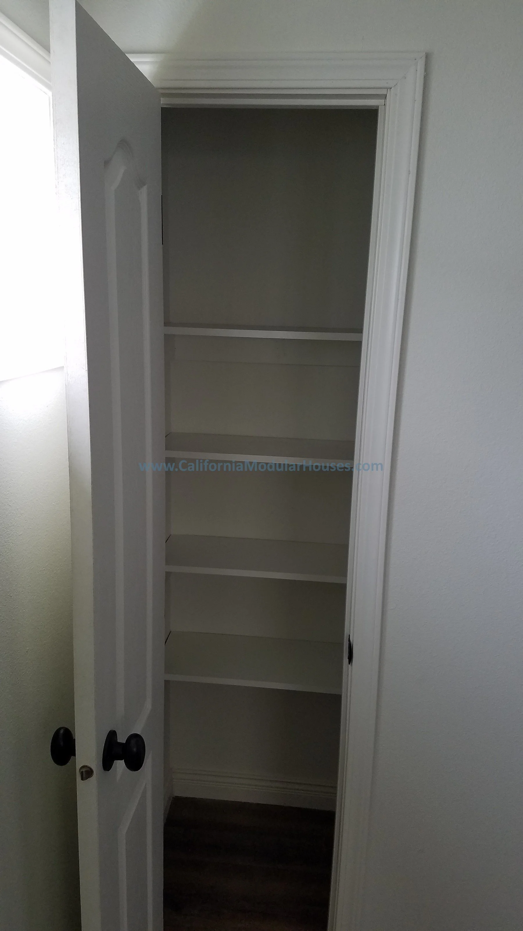 Empty white closet with shelves and black knobs. The closet door is open, revealing three shelves inside.  Prefab Modular Homes California, California Modular, California Modular Home, 