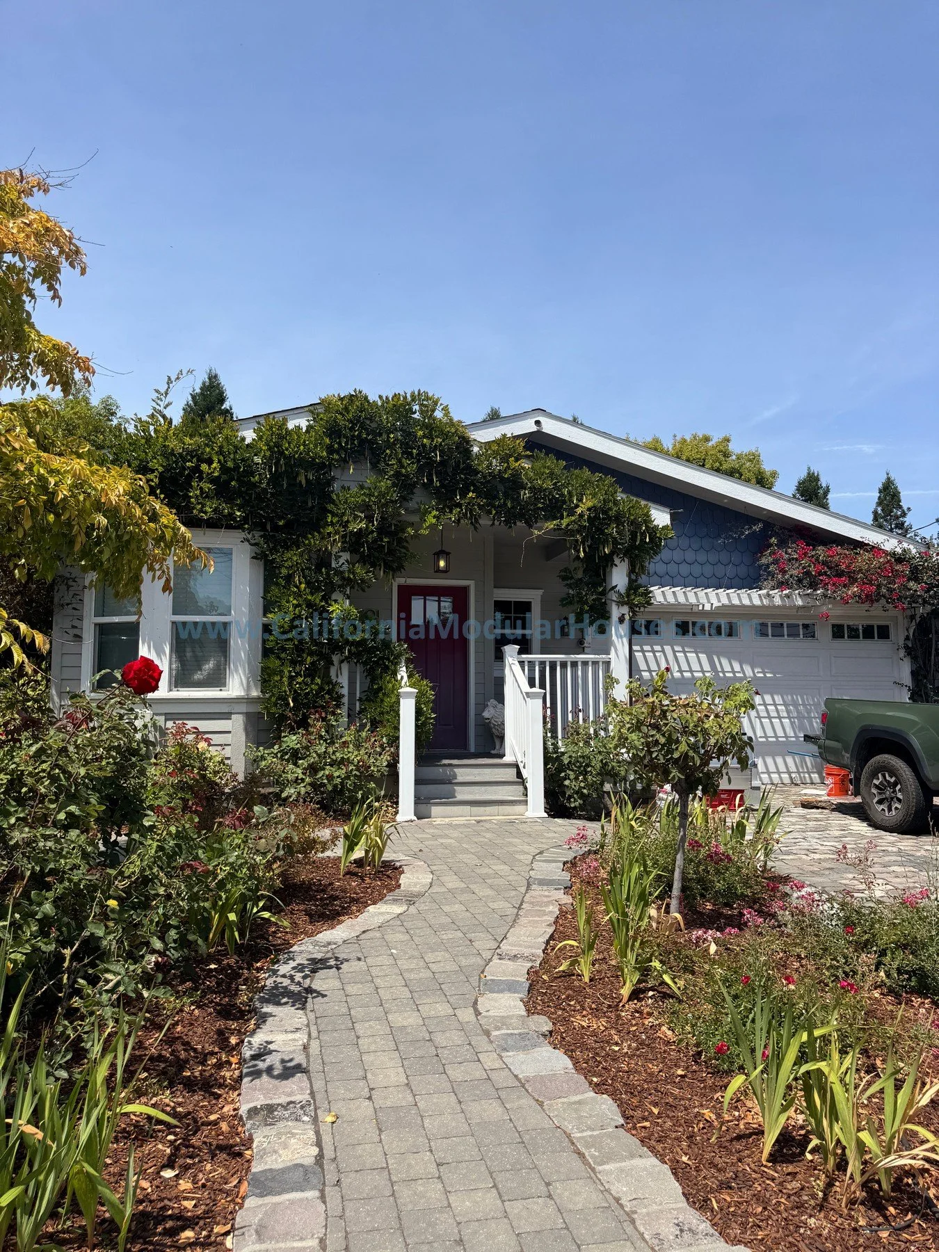 A cozy house with a red front door, a garden with blooming flowers, and a paved pathway leading to the entrance under a clear blue sky.