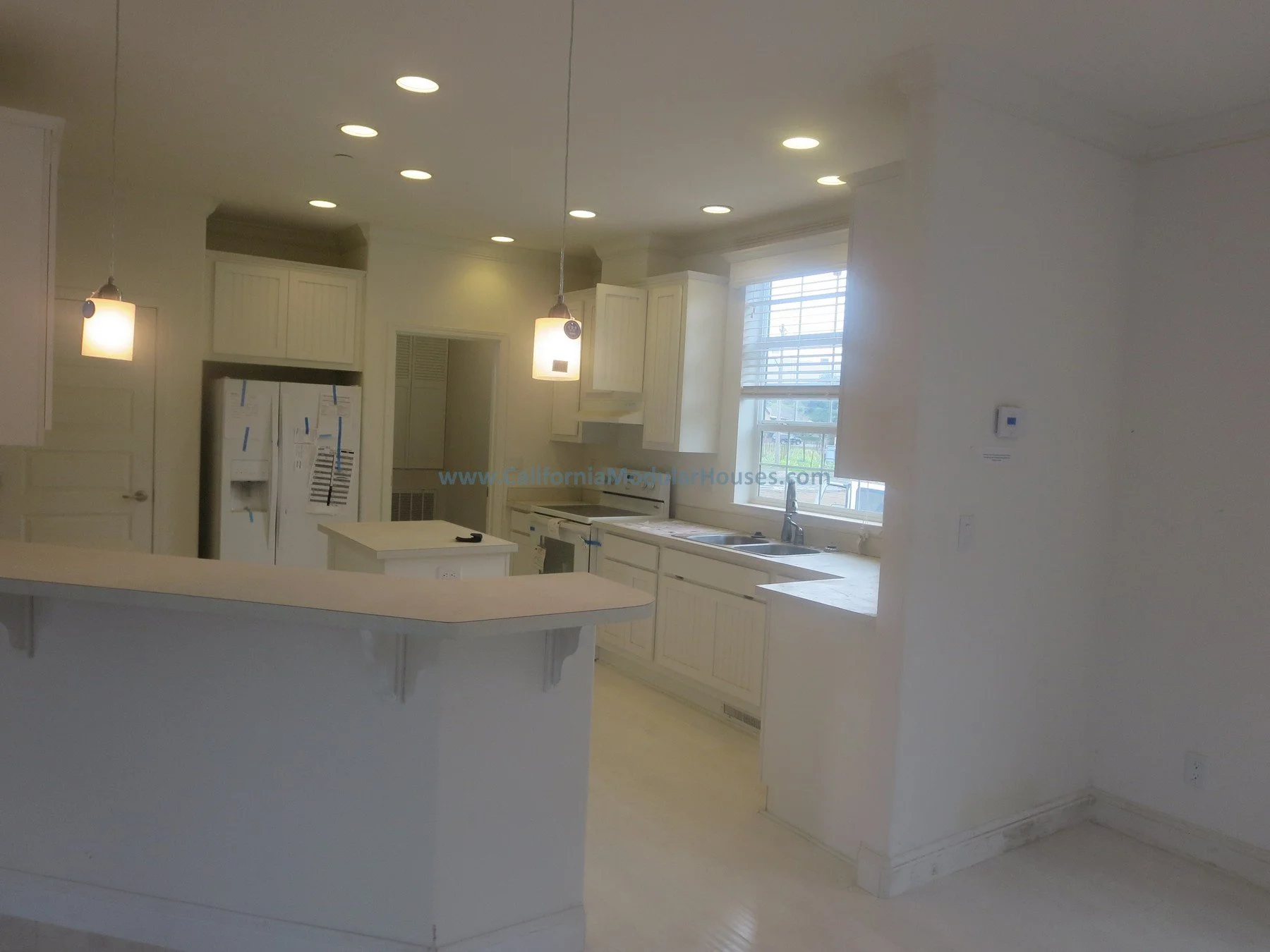 Empty kitchen with white cabinets, countertops, and a window, featuring pendant lights and a refrigerator. California Modular Home.  Single Family Residence.  Prefabricated.  Factory Built.  Napa, CA.  