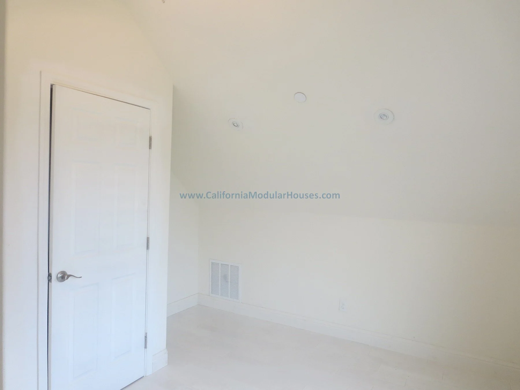 Empty white room with a closed door, three ceiling vents, a wall vent, and a power outlet, with light-colored flooring.  Napa, CA.  California Modular House. California modular home.  Prefab.  Modulars.  