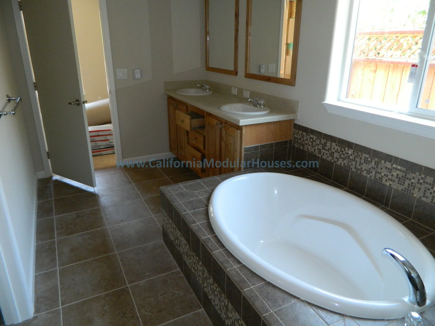 Bathroom with a corner bathtub, dual sinks with wooden cabinets, large window with a wooden fence outside, and tile flooring.