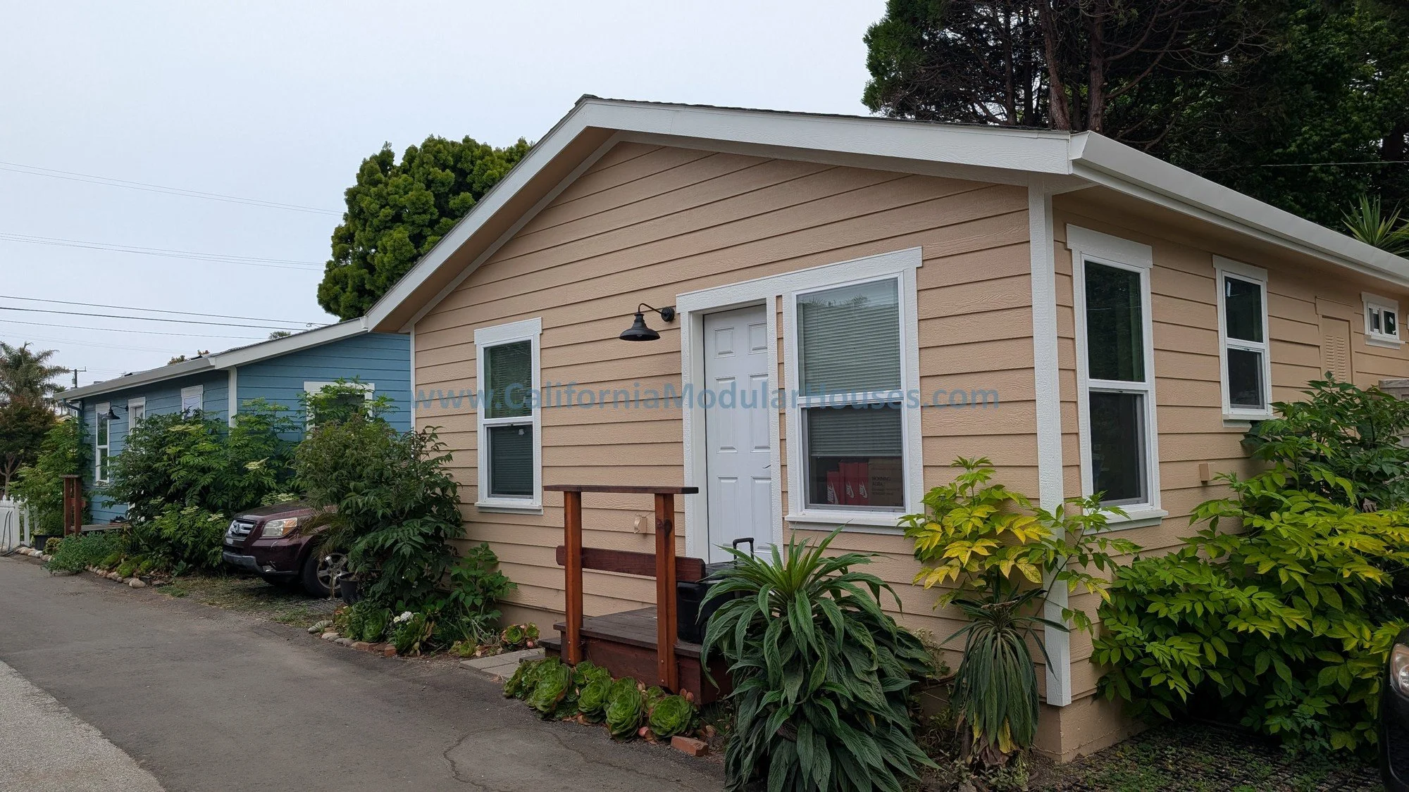 A single-story house with beige and blue siding, white trim, and multiple windows, surrounded by greenery and plants, with a small wooden porch and a parked car nearby.