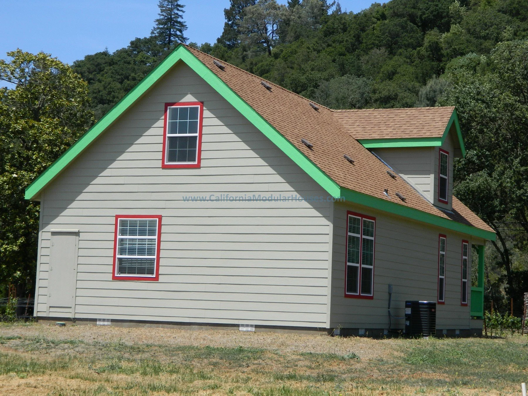 A two-story house with beige siding, red window frames, and a brown shingle roof with small vents. Green trim accents the roofline and porch area. The house is on a grassy lot with trees and hills in the background.