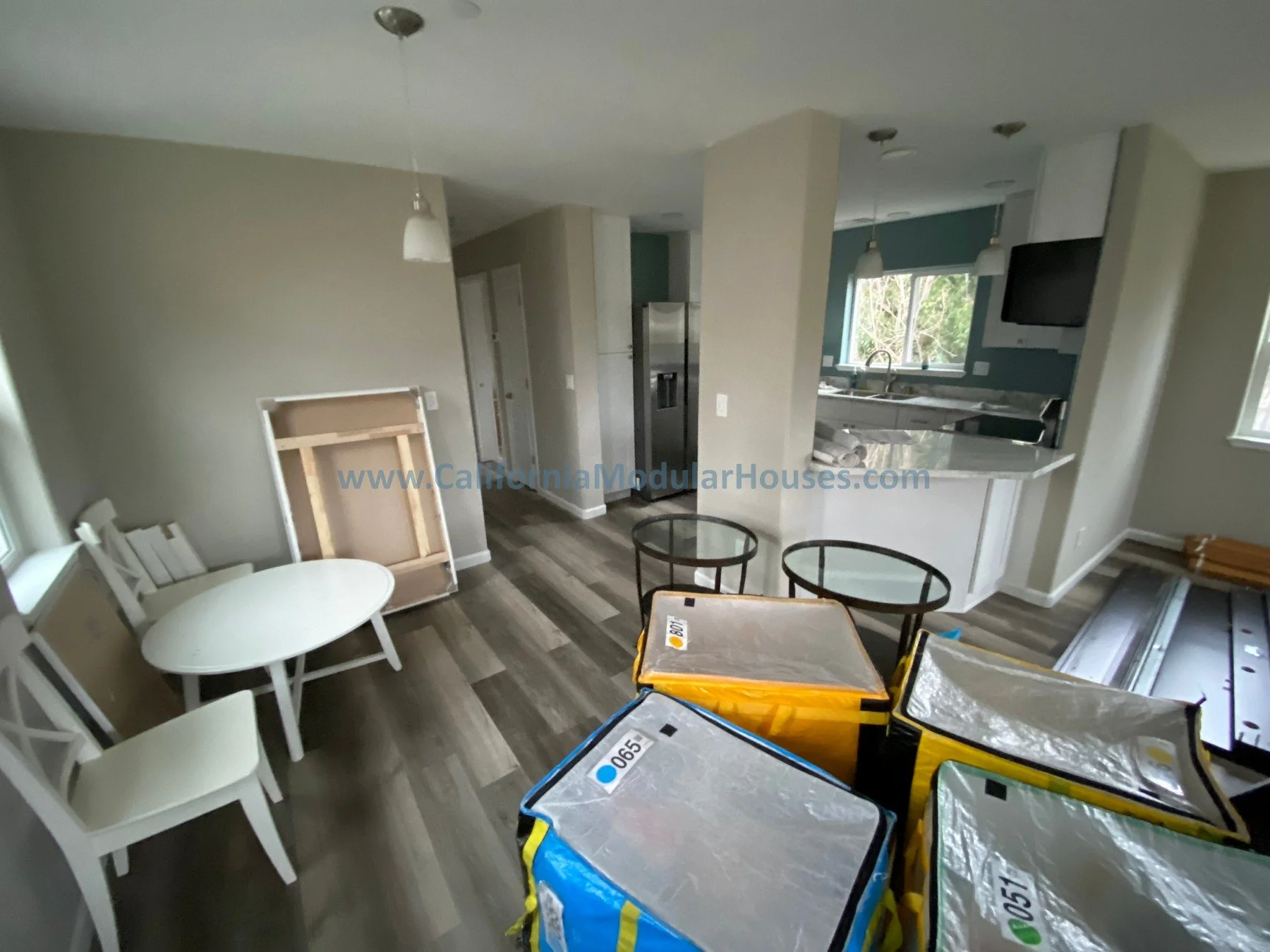 California modular.   Modular CA Living room under renovation with stacked furniture, a small dining table with chairs, a kitchen with an island, window, and a wall-mounted TV.