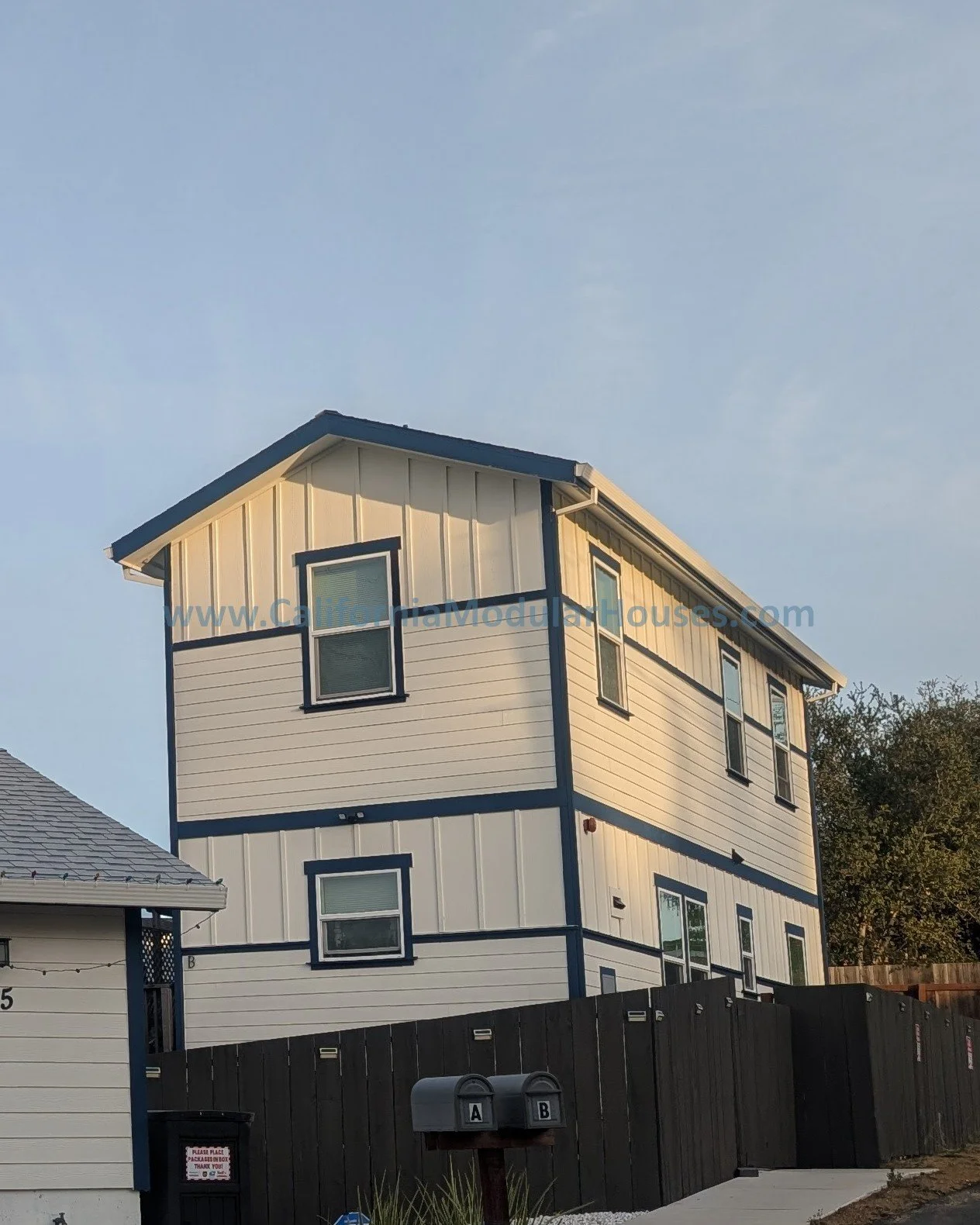 Three-story modern tiny house with white and blue exterior, multiple windows, and a sloped roof, situated behind a black privacy fence.  Accessory Dwelling Unit Prefab Modular Home for Contra Costa County, CA.  Modular ADU California.