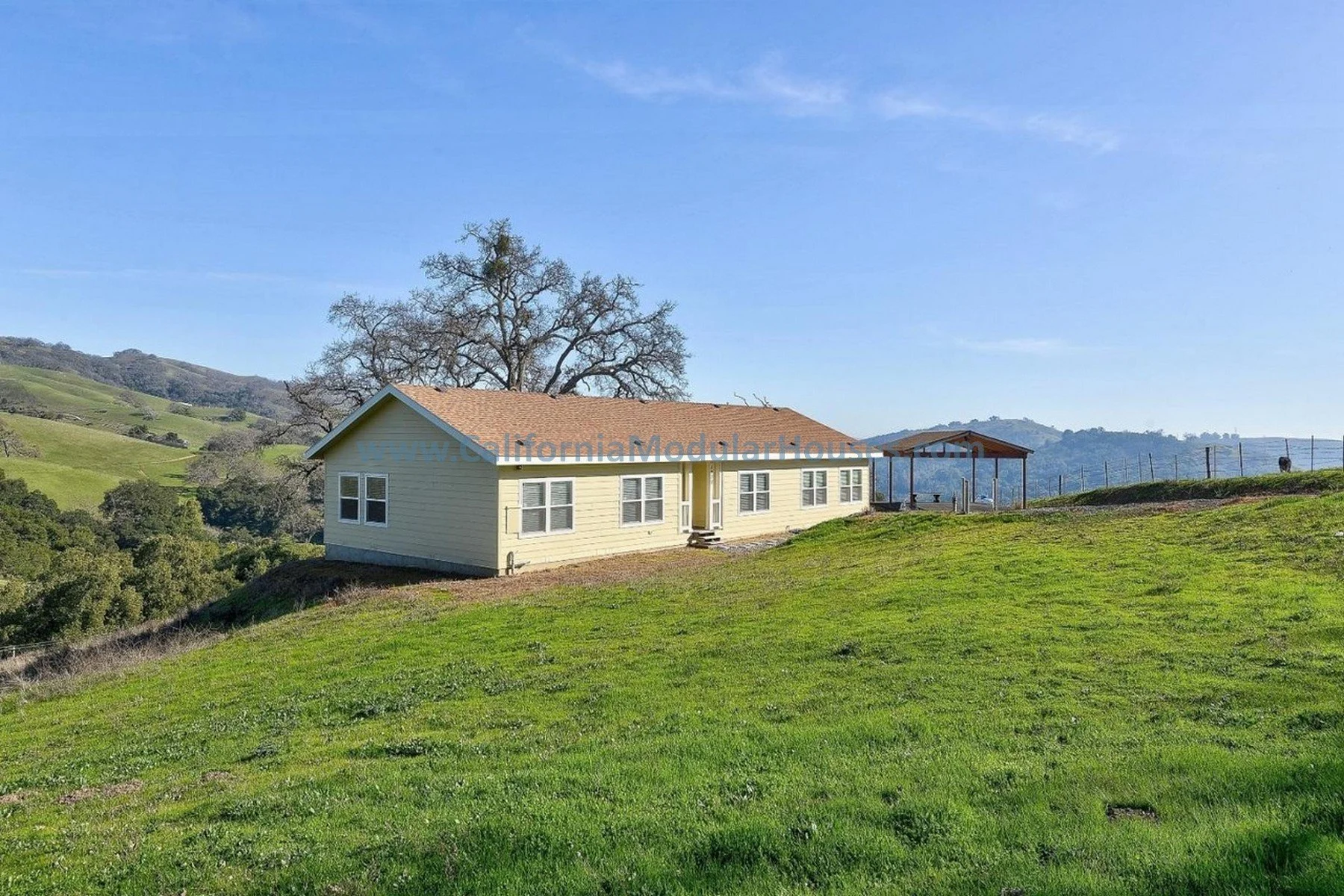 A single-story beige house with white trim sits on a grassy hill under a clear blue sky, with rolling hills and trees in the background.  Bay Area Prefab Homes, Modular Home on a Sloped Property. 