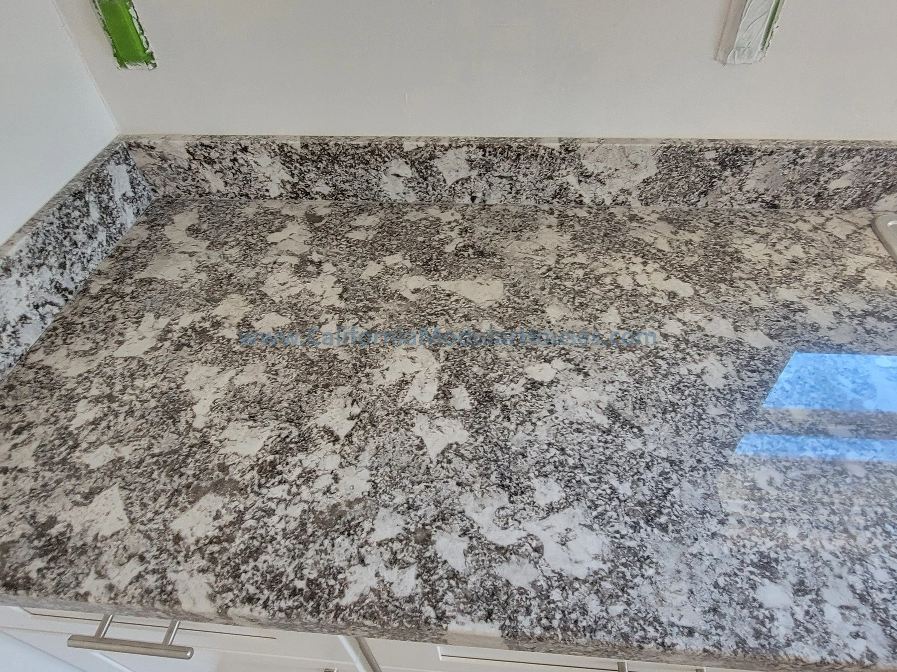 Close-up view of a brown and white speckled granite countertop in a kitchen, with a workspace near the wall, some electrical outlets, and a reflection of window blinds on the surface.