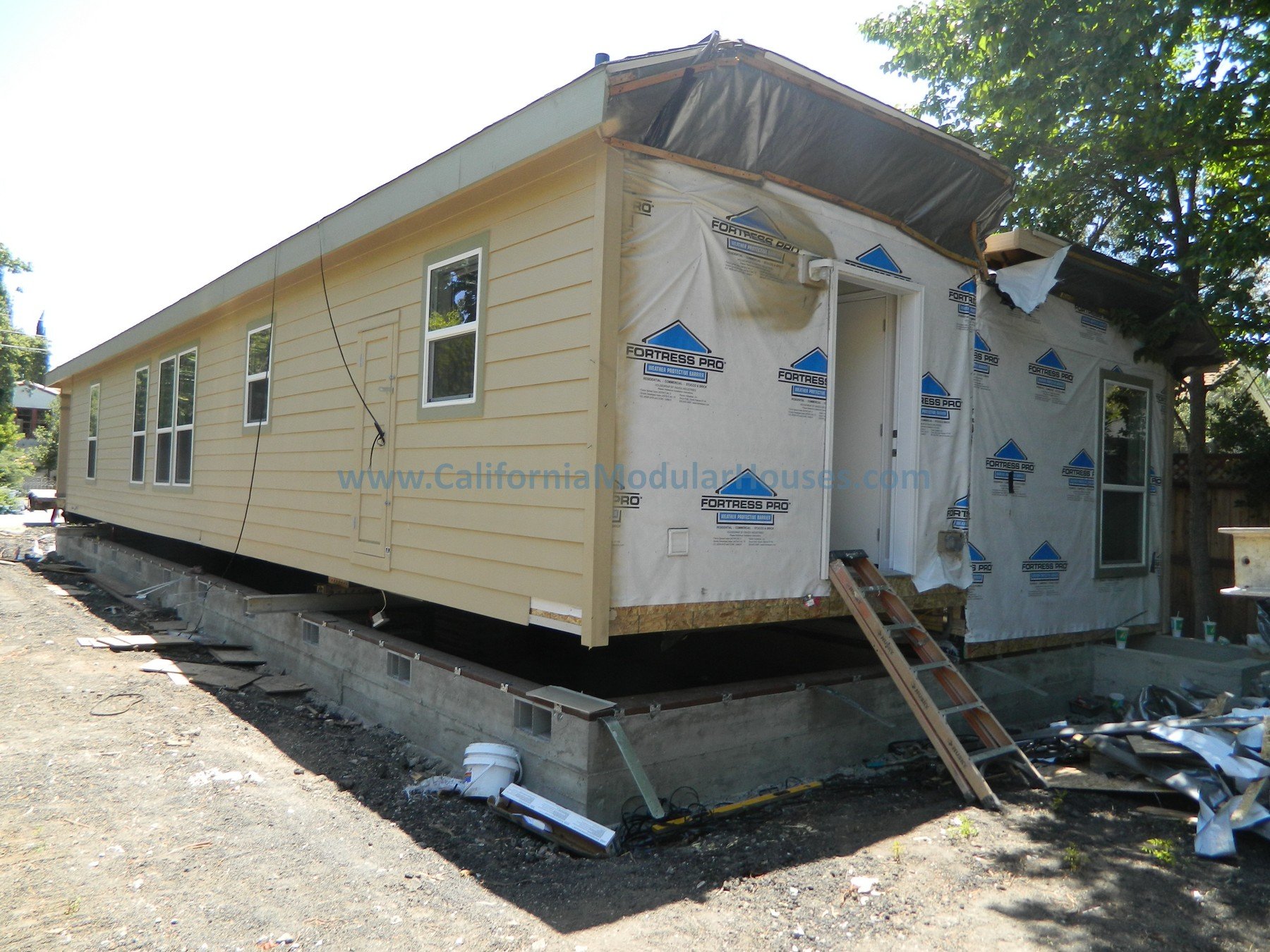 A house under construction with yellow siding, multiple windows, and a concrete foundation. Part of the house has exterior wall sheathing and temporary ladder, with construction materials and tools scattered around.