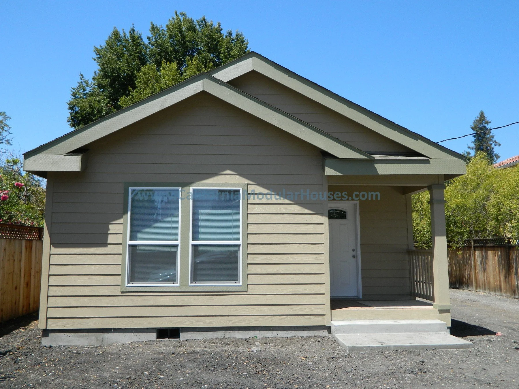 A small beige house with a peaked roof and white window frames, surrounded by a wooden fence and green trees, under a clear blue sky.