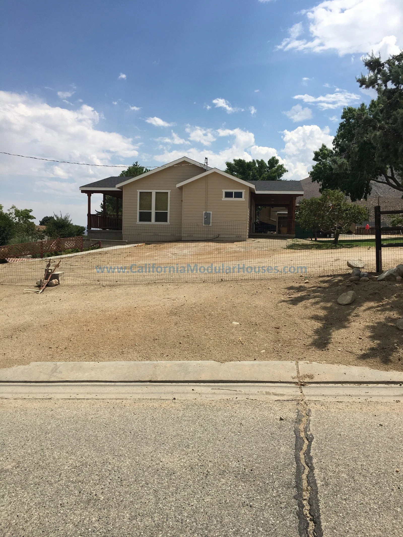 A new beige house with white windows and dark roofing, surrounded by a sandy yard and a chain-link fence, under a partly cloudy sky.