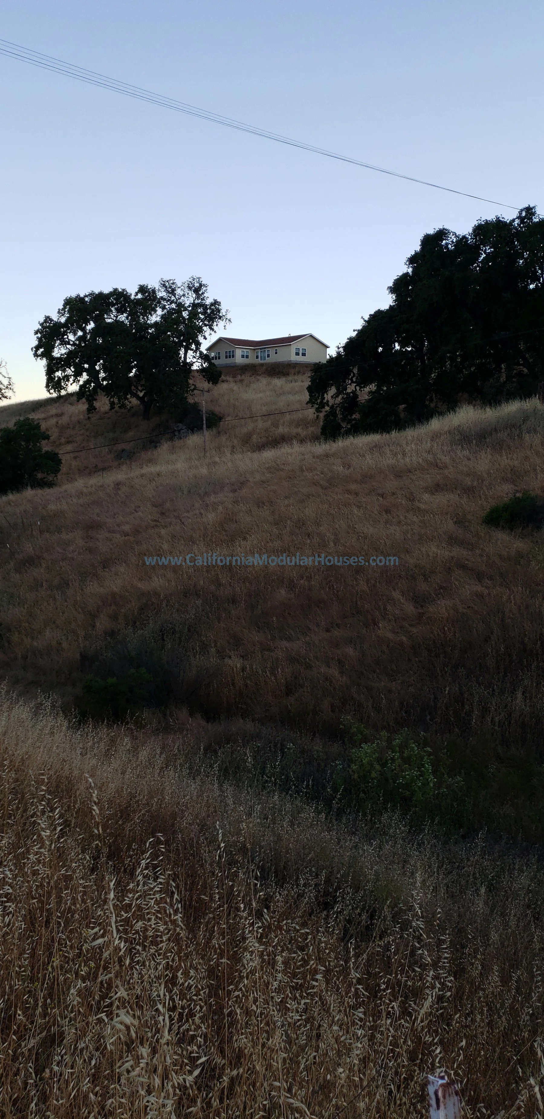 A house on a hill surrounded by dry grass and trees under a clear blue sky, with power lines overhead.  Bay Area Modular Home, California Modular Homes, Prefab Modular Homes California.