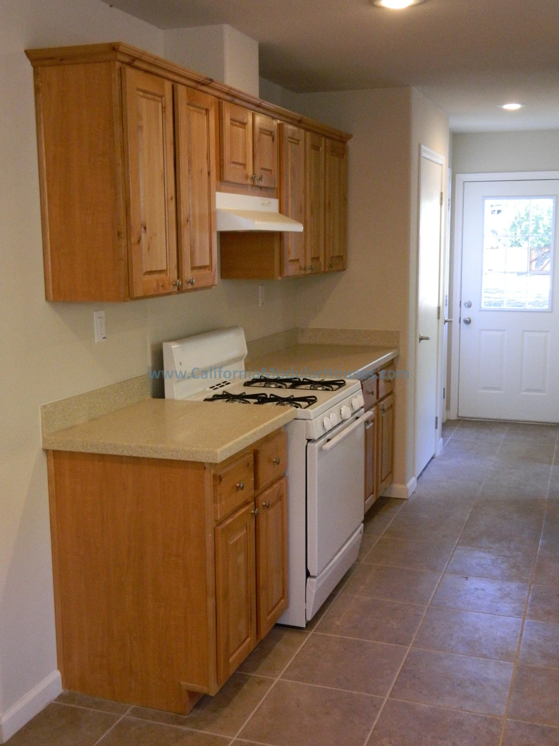 Kitchen with wooden cabinets, beige countertops, and a white stove. The floor is tiled, and there is a door leading outside in the background.