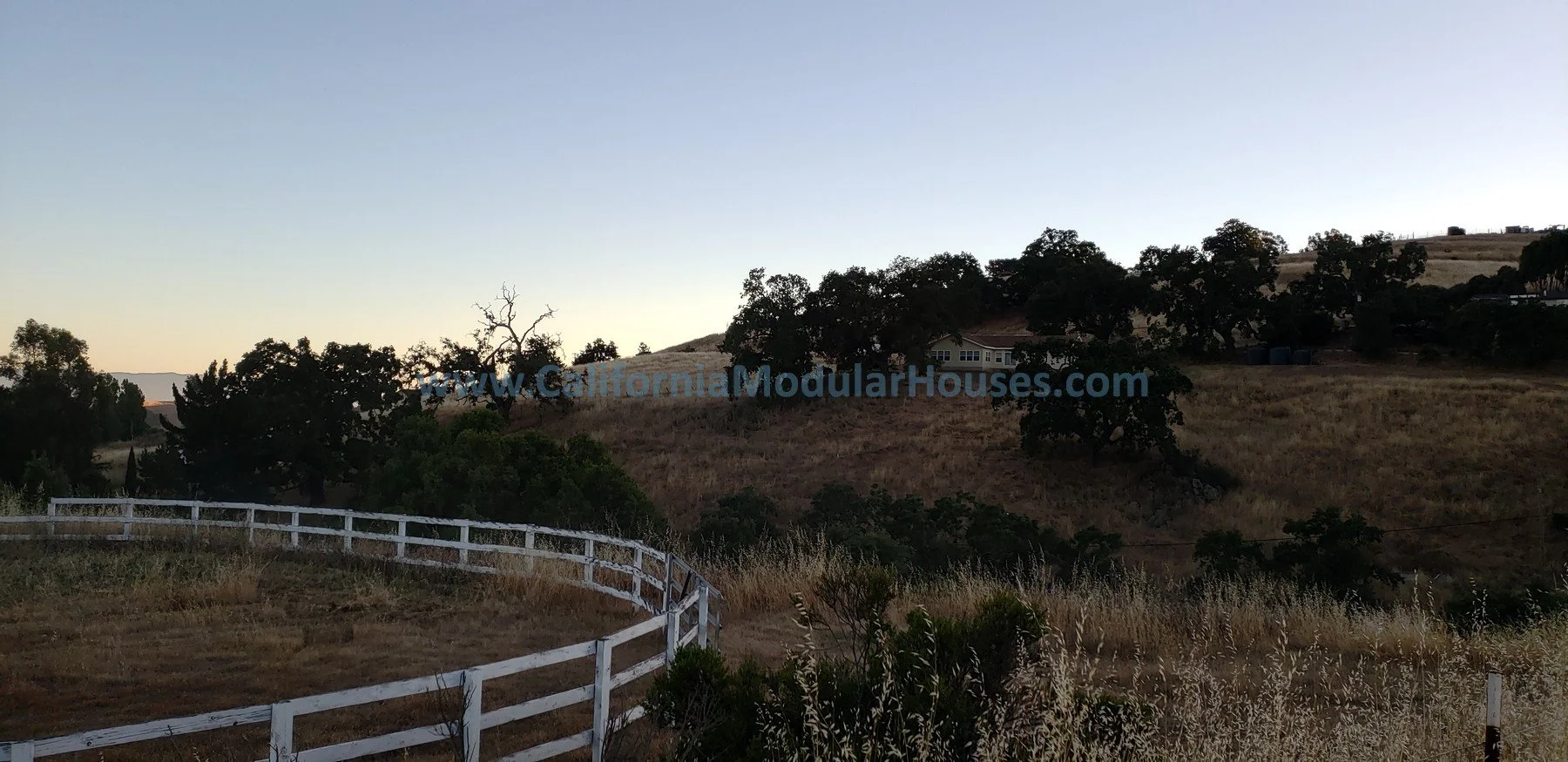 Hilly landscape with dry grass, scattered trees, a white wooden fence in the foreground, and a house on the hillside at sunset. California Modular Houses, Modular Home on a Sloped Property. 
