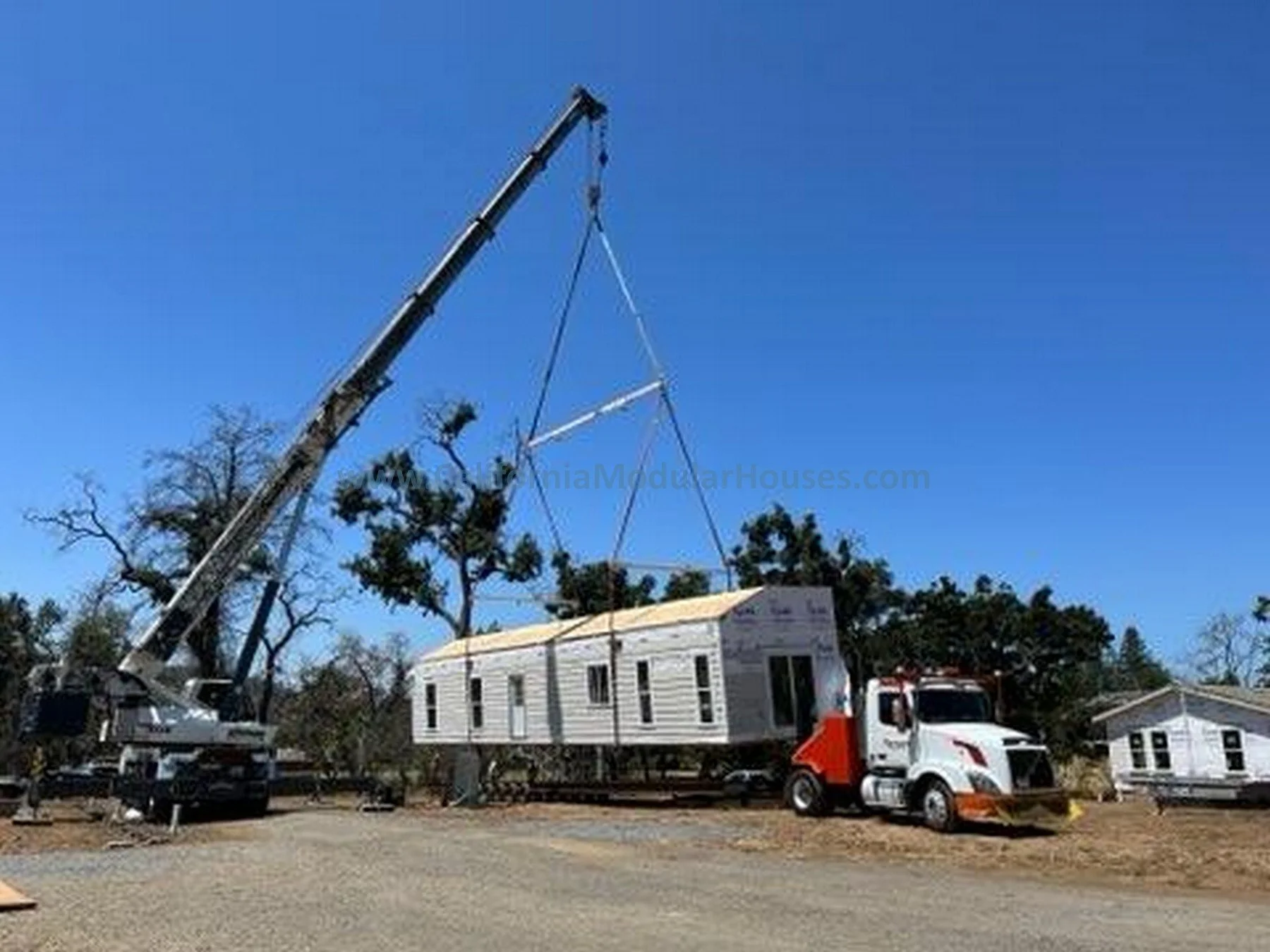 A crane lifting a mobile home onto a truck at a construction site with trees and other similar houses in the background under a clear blue sky.
