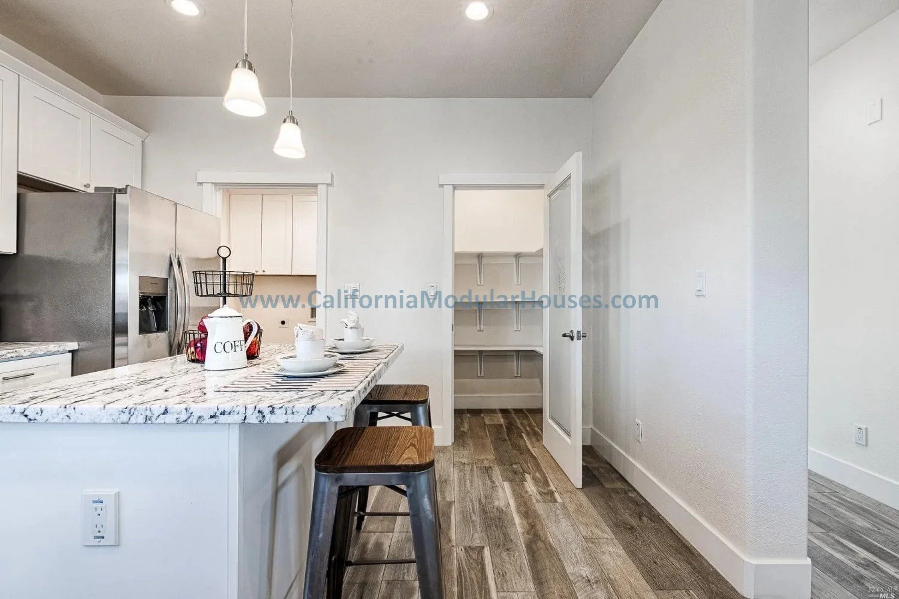 Kitchen with white cabinets, stainless steel refrigerator, marble countertop island, two bar stools, and doorway leading to pantry with built-in shelves.
