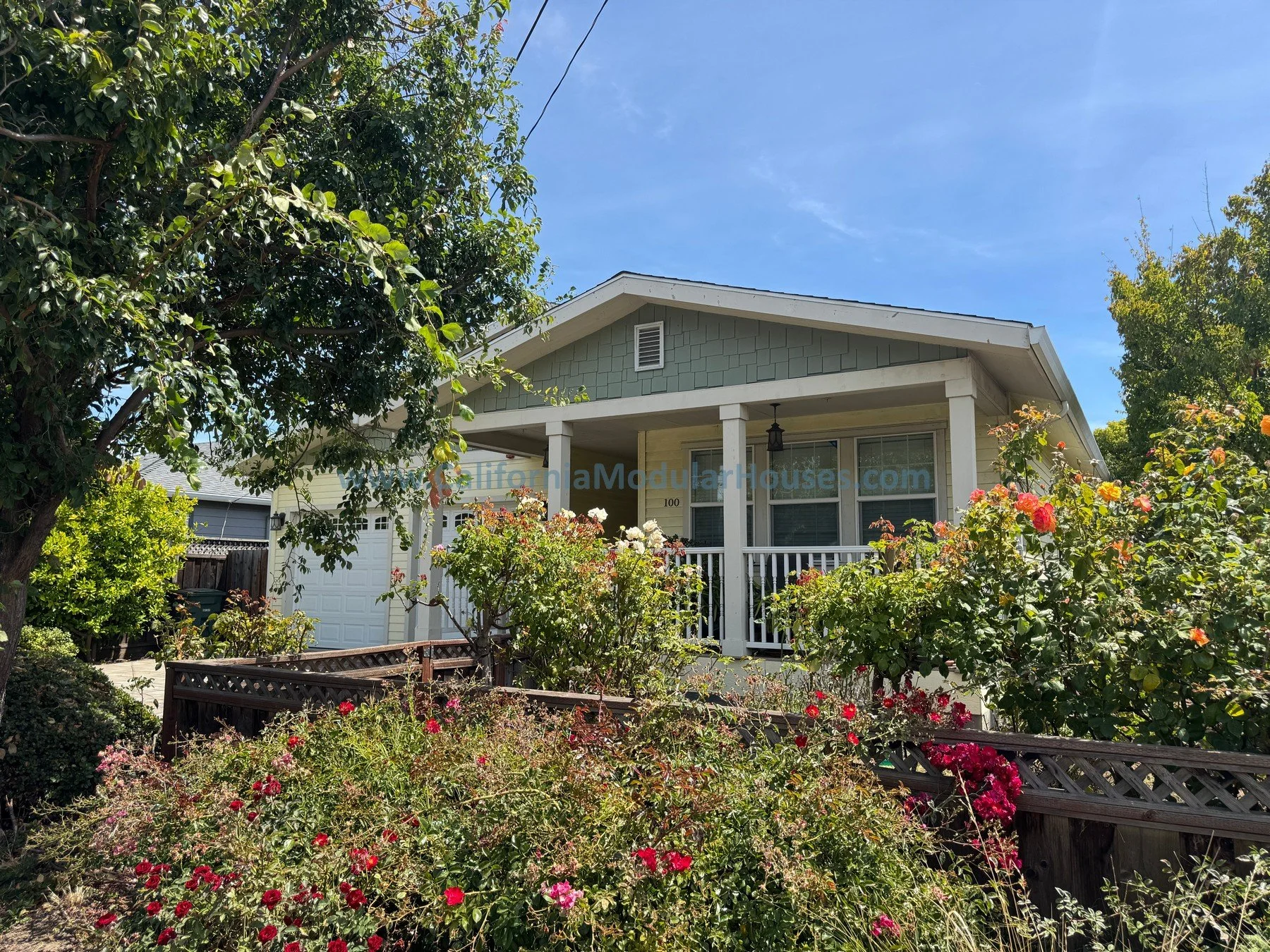 A house with a front porch surrounded by a lush garden of colorful flowers and green foliage, under a clear blue sky.