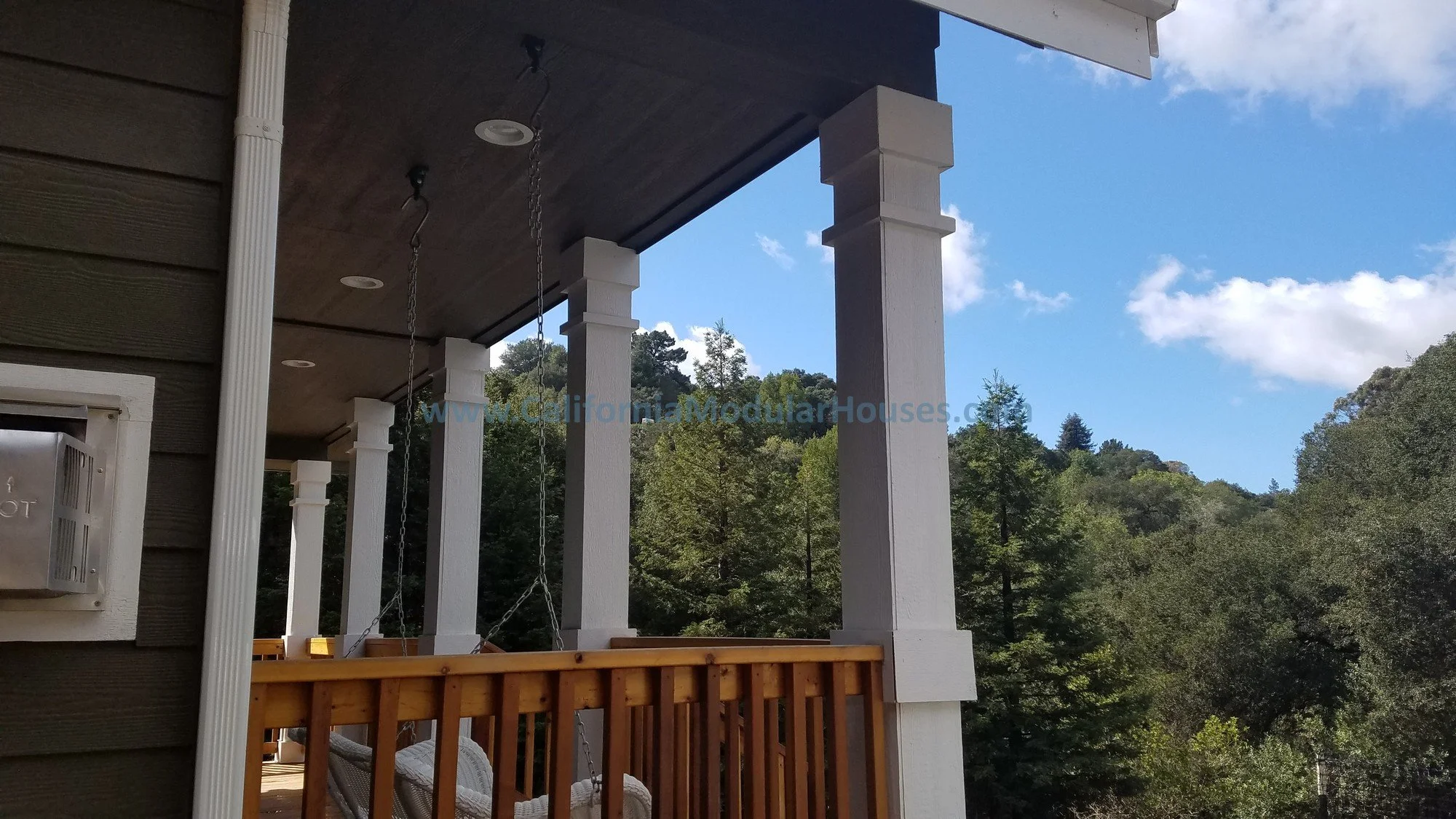 View of a house porch with white columns, a dark ceiling, and hanging swing chains, overlooking a green wooded hillside under a partly cloudy blue sky.  Factory Built Housing.  