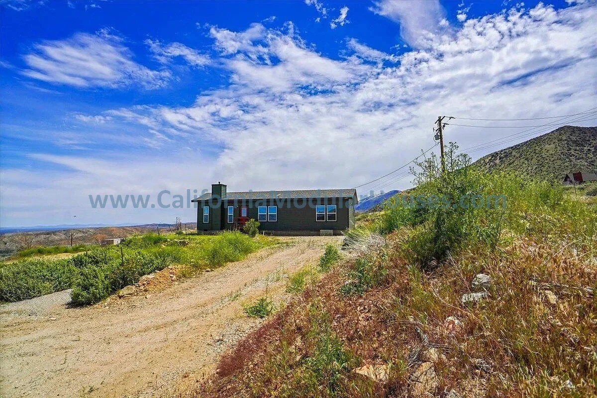 A small house with six windows, painted dark gray, is situated in a semi-arid landscape with sparse green bushes and a gravel driveway. The sky is partly cloudy with streaks of white clouds across a blue sky, and mountains or hills are visible in the
