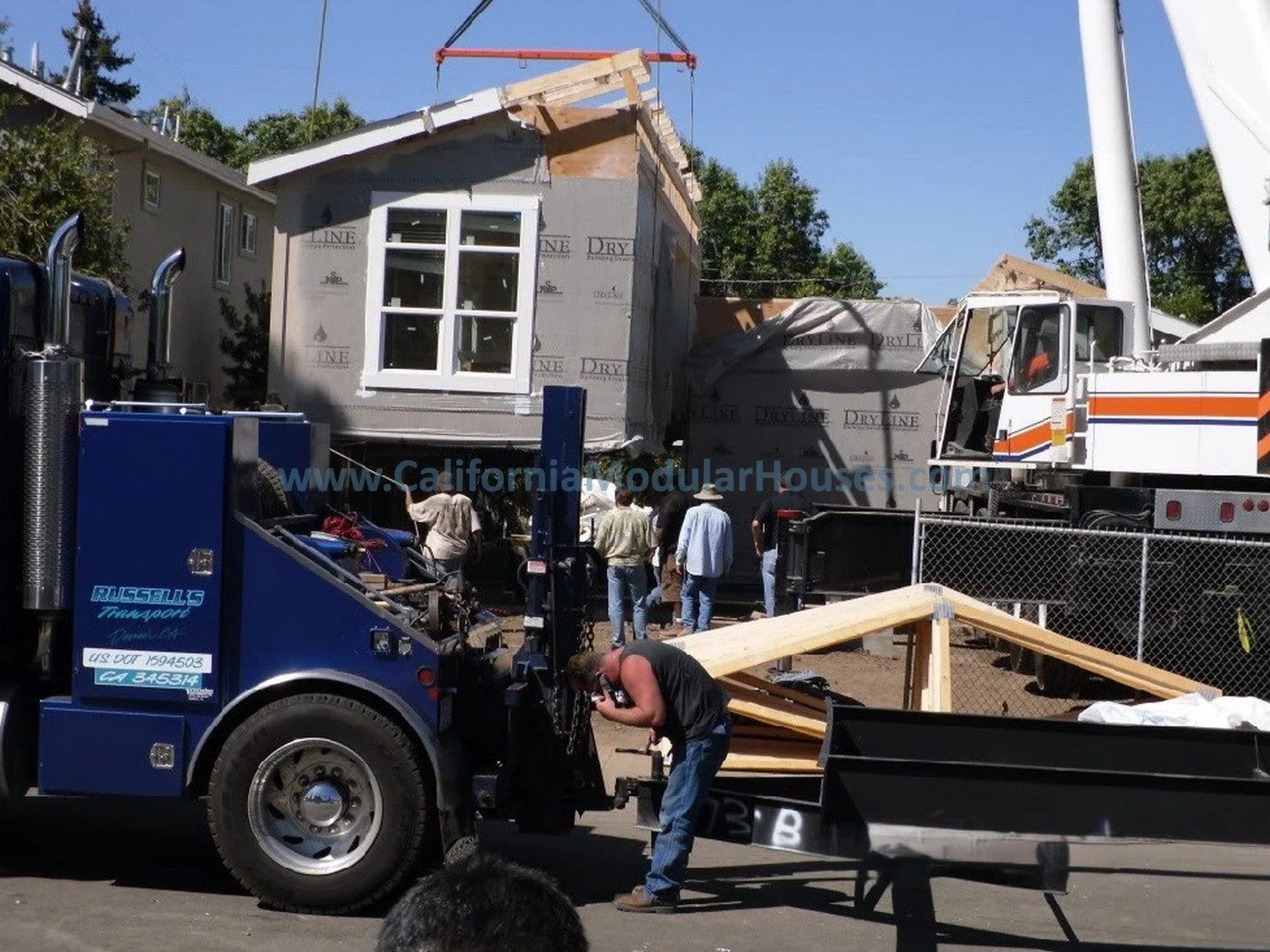 A prefab modular house section is being lifted by a large crane onto a flatbed truck at a construction site, with several workers and onlookers present, and a wooden ramp leading up to the truck.