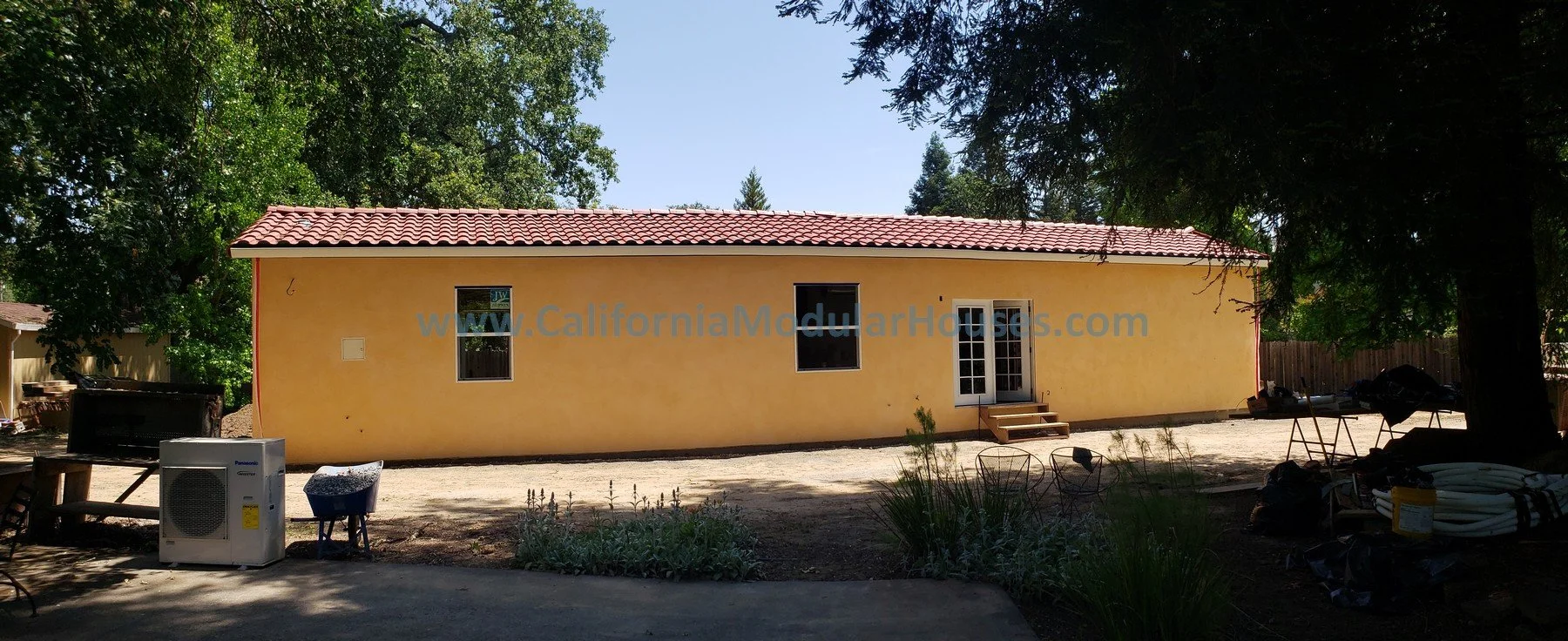 A yellow house with a red tiled roof, white framed windows, a glass door with a small set of stairs, surrounded by trees and natural ground.  2 bedroom, 2 bathroom modular ADU from the exterior.   