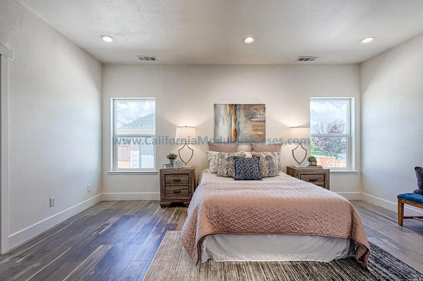 Bedroom with a bed, two nightstands with lamps, and two windows showing a cloudy sky and neighboring roofs.
