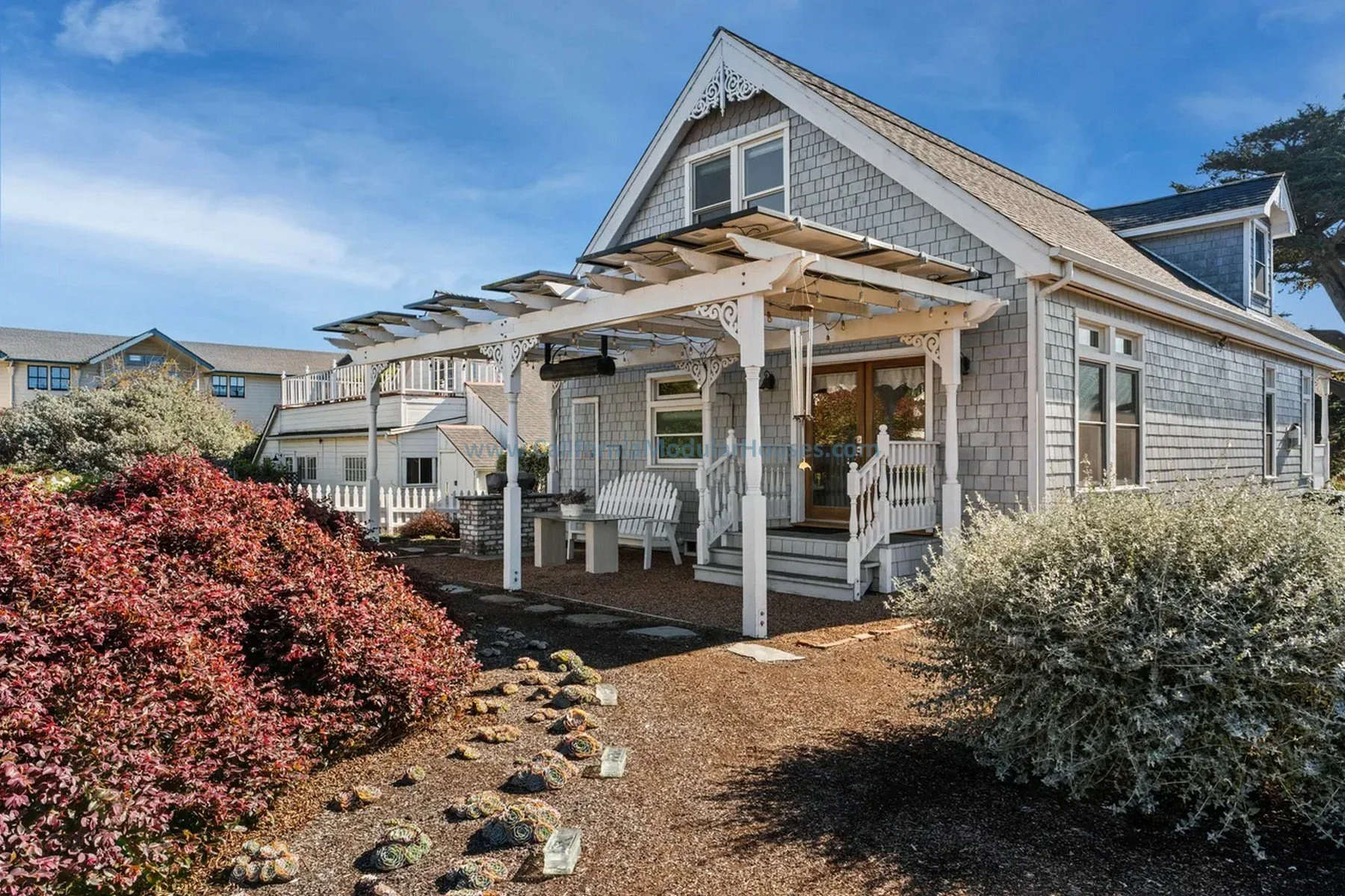 Historical design. Mendocino City, CA. Factory Built Home.  Mendocino County, California. A charming seaside house with a covered front porch, white decorative trim, and shingle siding. The pathway leading to the porch is lined with rocks. 