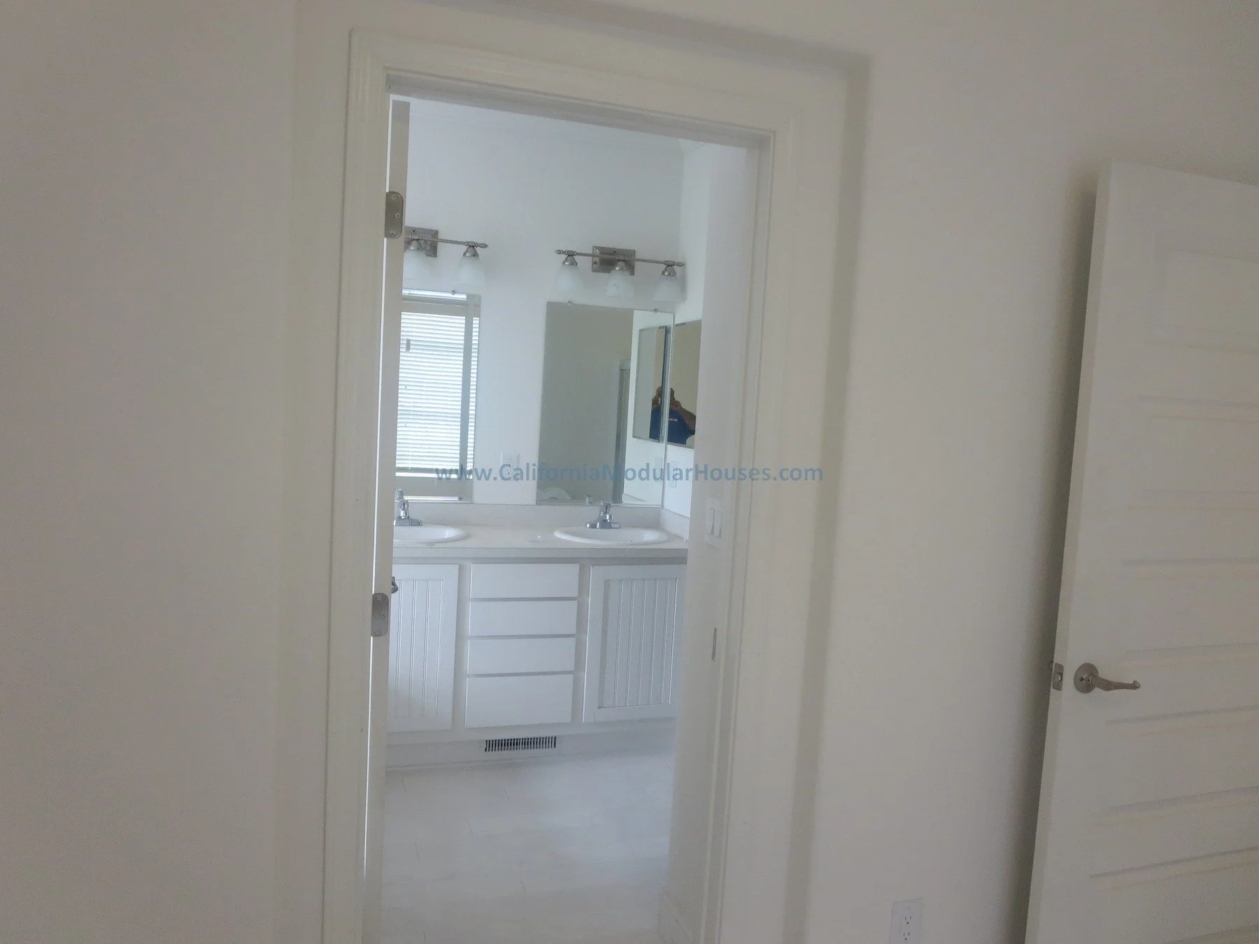 Cape Cod.  Napa County, CA. Modular. Pre-fab Modular.   Modulars for Napa.  View of a bathroom with a double sink vanity, mirror, and light fixtures, seen through a doorway in a bright, white-walled room.  California.  