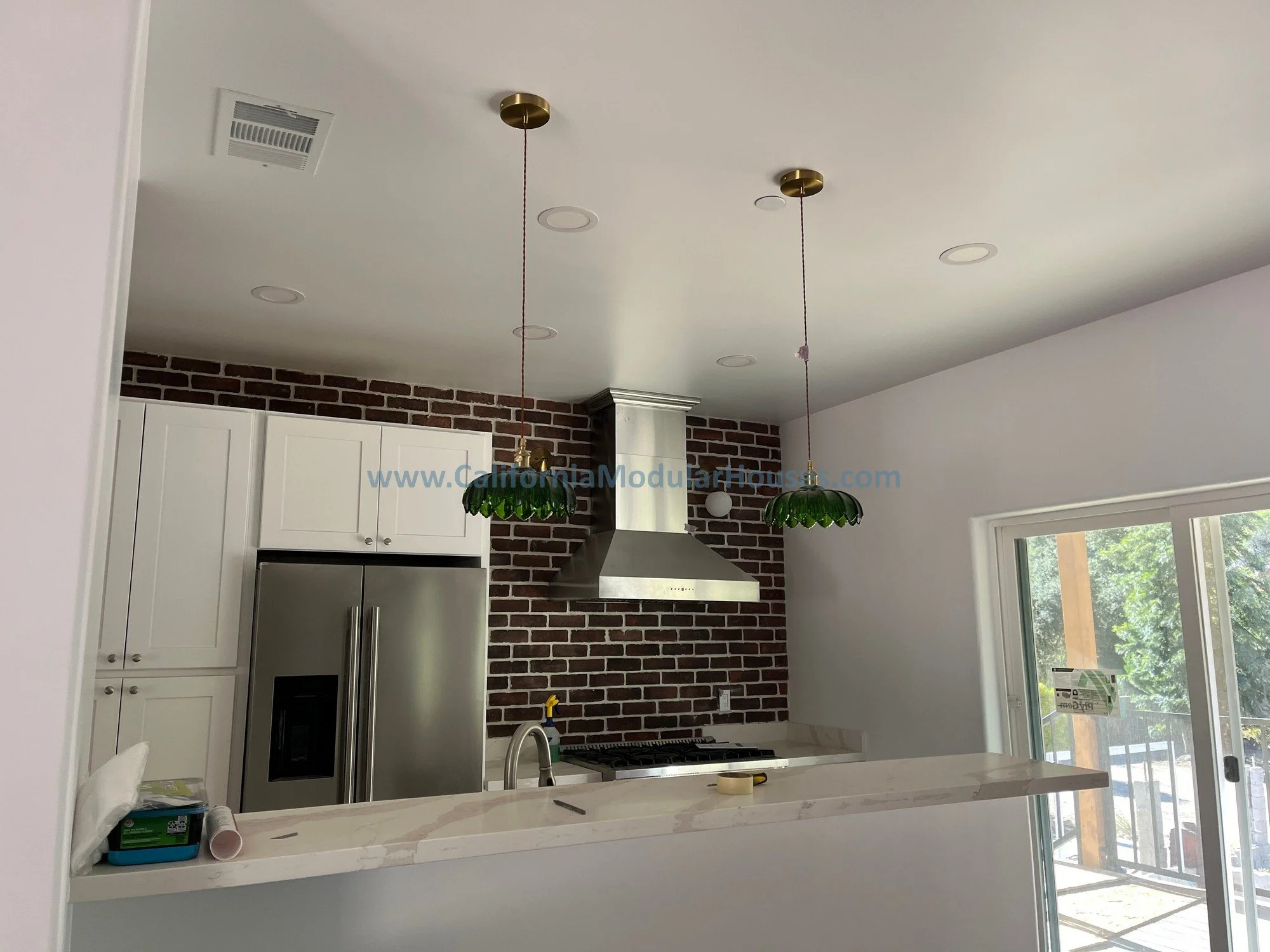 Modern kitchen with white cabinets, stainless steel refrigerator, brick wall, stainless steel range hood, black stove, white marble countertops, sliding glass door, and green pendant lights.