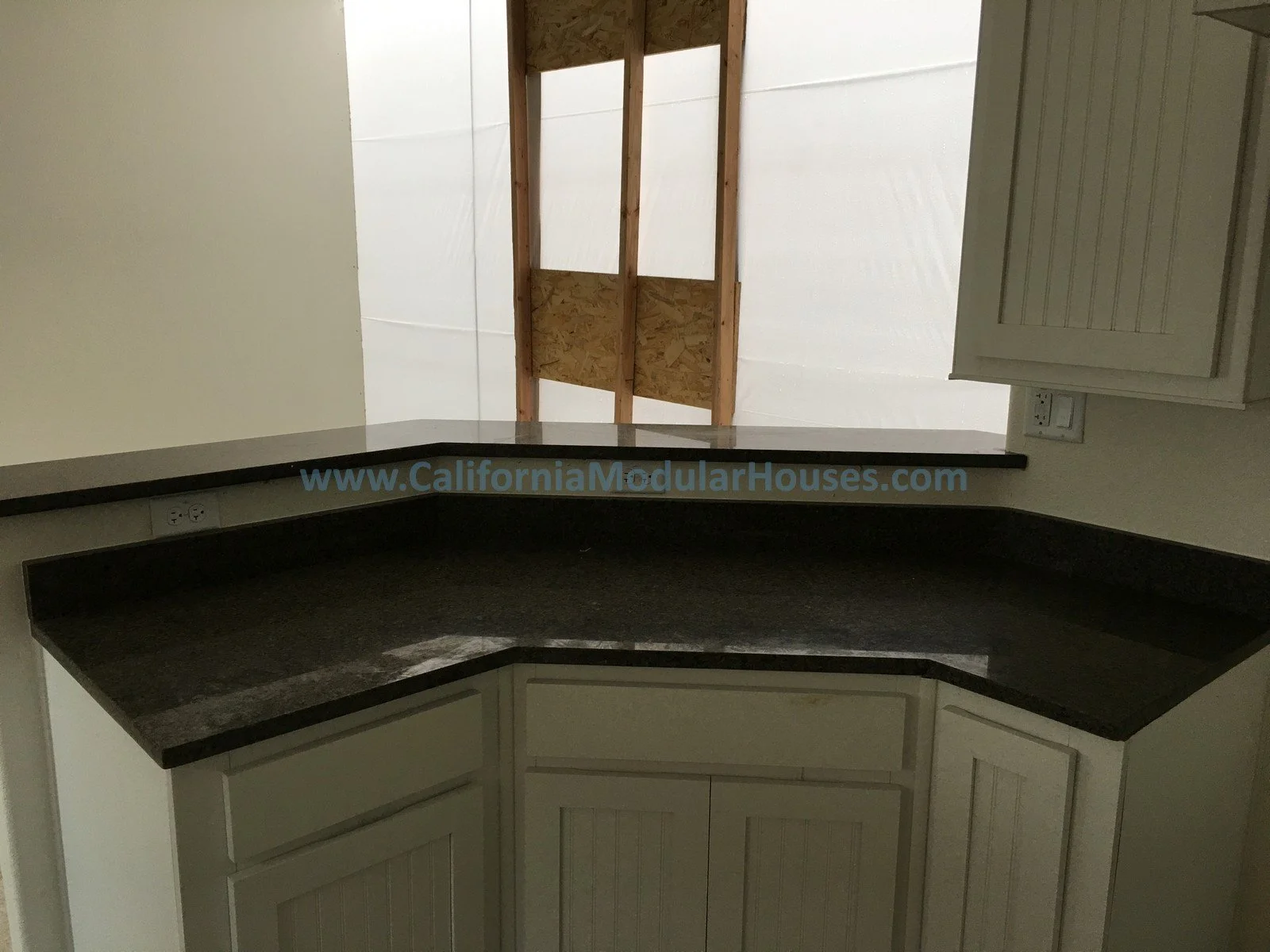 Interior view of a kitchen with a black quart countertop, white wooden cabinets, and an open area with and a passthrough shown for this modular home.  