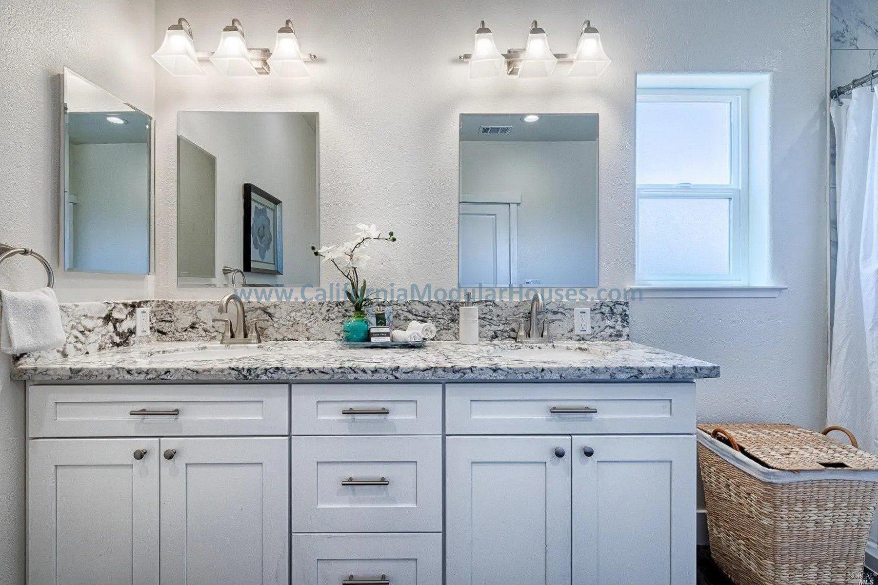 Bathroom vanity with two sinks, granite countertop, two mirrors, and a small window. Decor includes a flower in a turquoise vase, a tray with toiletries, and a soap dispenser. There is a wicker laundry basket on the right side.