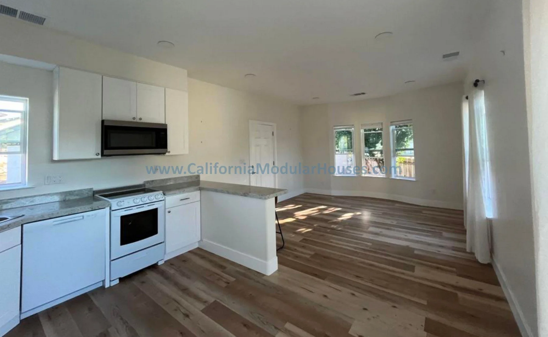 Interior view of a kitchen and living area in a house, featuring white cabinets, a white stove, microwave, and dishwasher, with wood flooring and multiple windows allowing natural light, with walls painted in a light color.  Modular Home CA