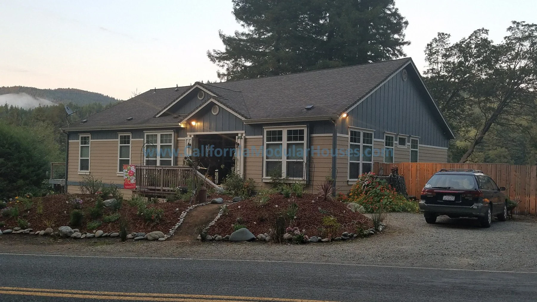 A house with siding, multiple windows, and a front porch with steps. The scene is set in a rural or semi-rural area with trees and hills in the background.  Crescent City Modular Home, Pre-Fabricated Homes.
