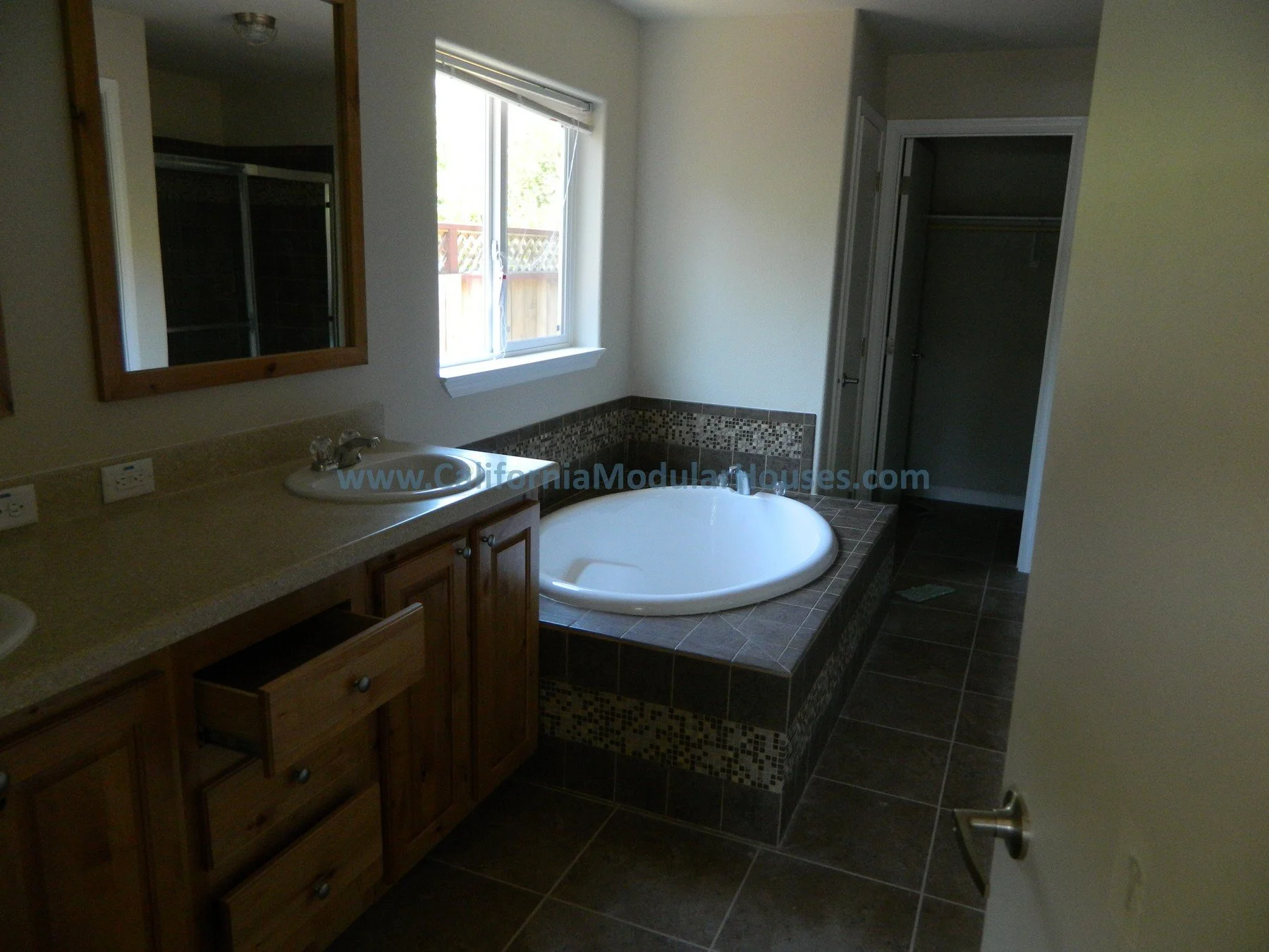 Bathroom with a wooden vanity, a mirror, a window, a corner bathtub surrounded by brown tiles with mosaic accents, and an open closet.