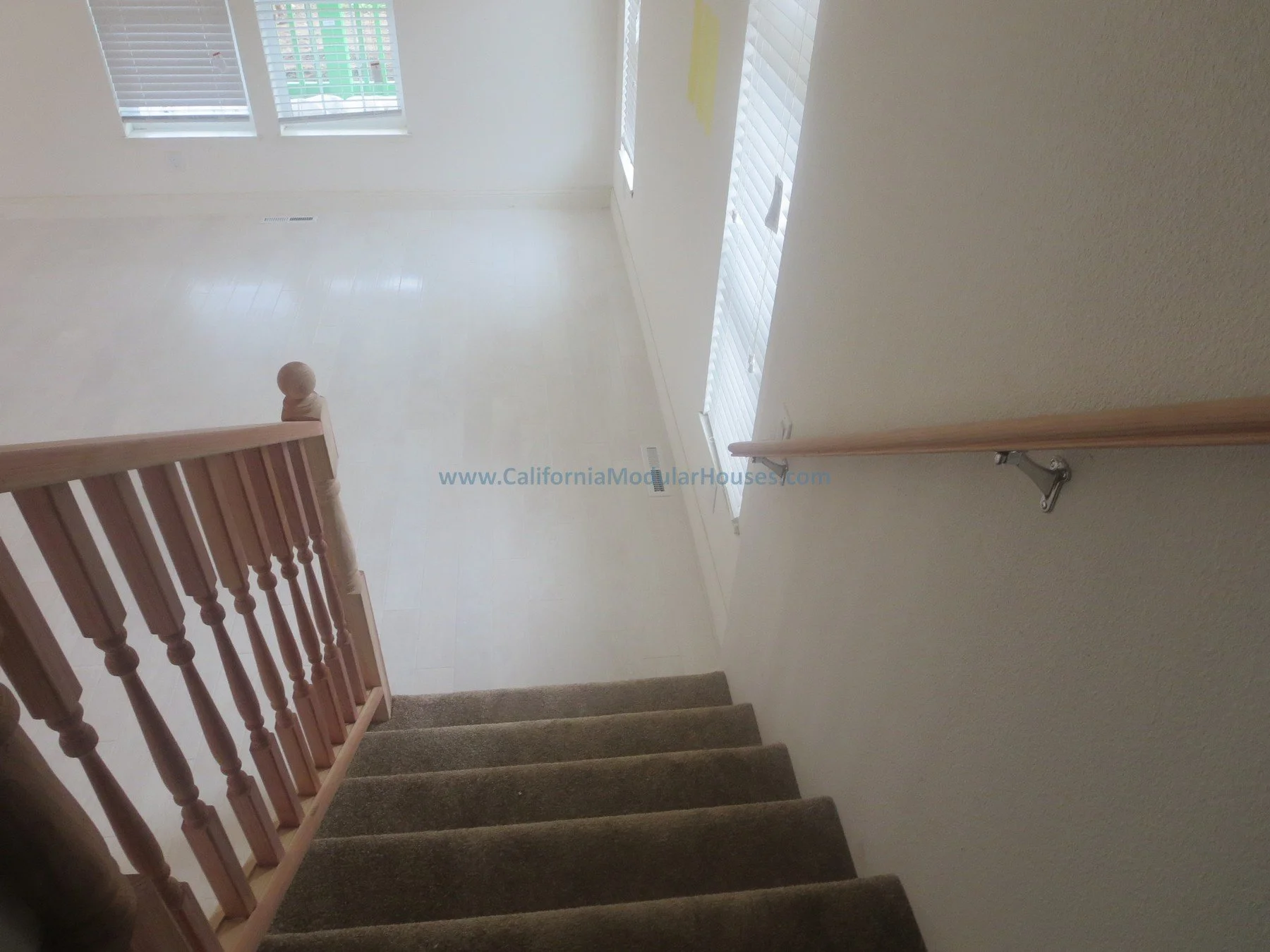 Interior view of a staircase leading down to a bright, open living space with three windows and white walls, with a wooden railing on the left side and stairs.  Prefab.  Napa, CA.  California Modular House. California modular home.