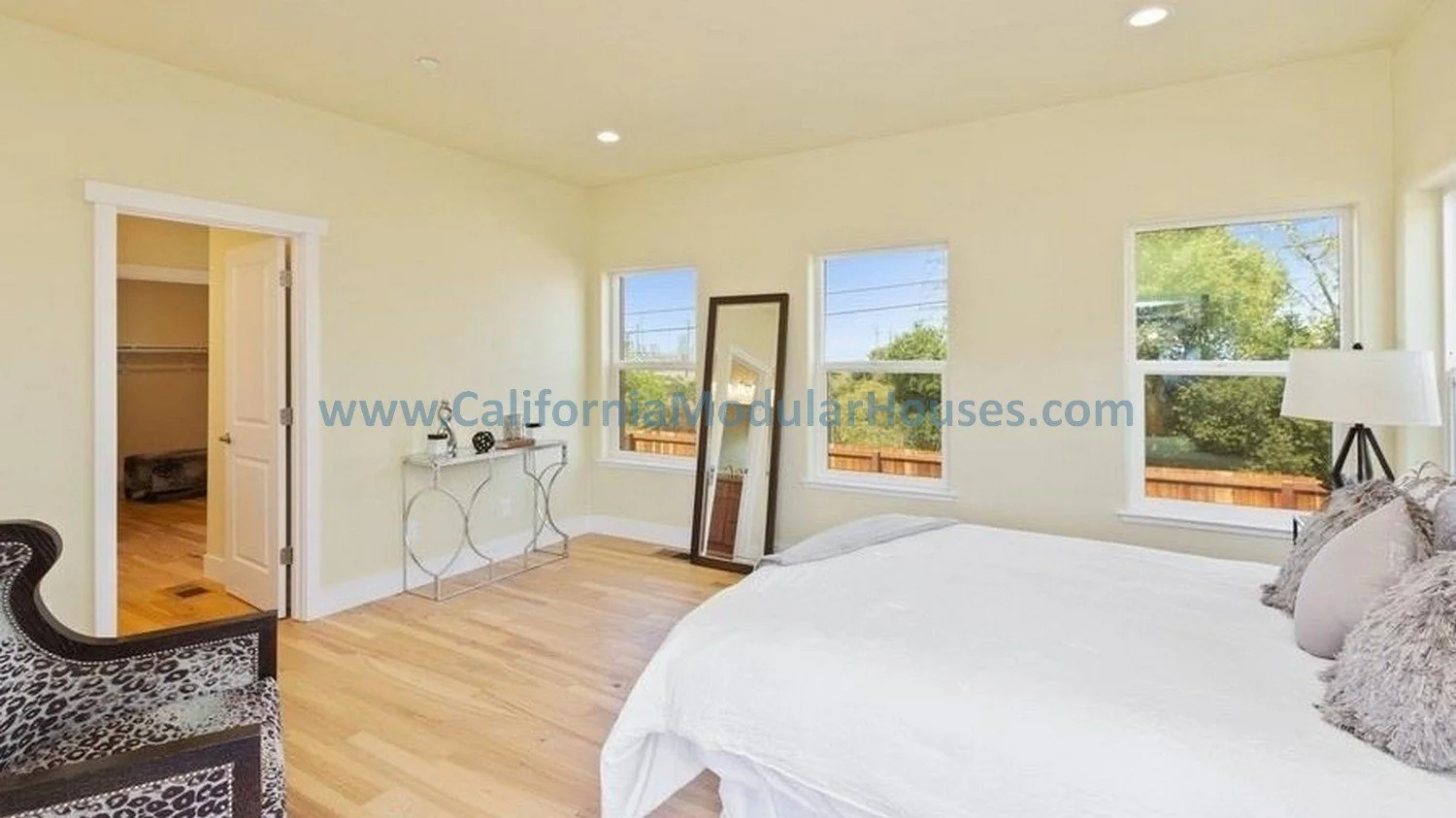 Bright bedroom with three windows, a large bed, a standing mirror, a floor lamp, and an open closet door.  California modular home.  prefab.