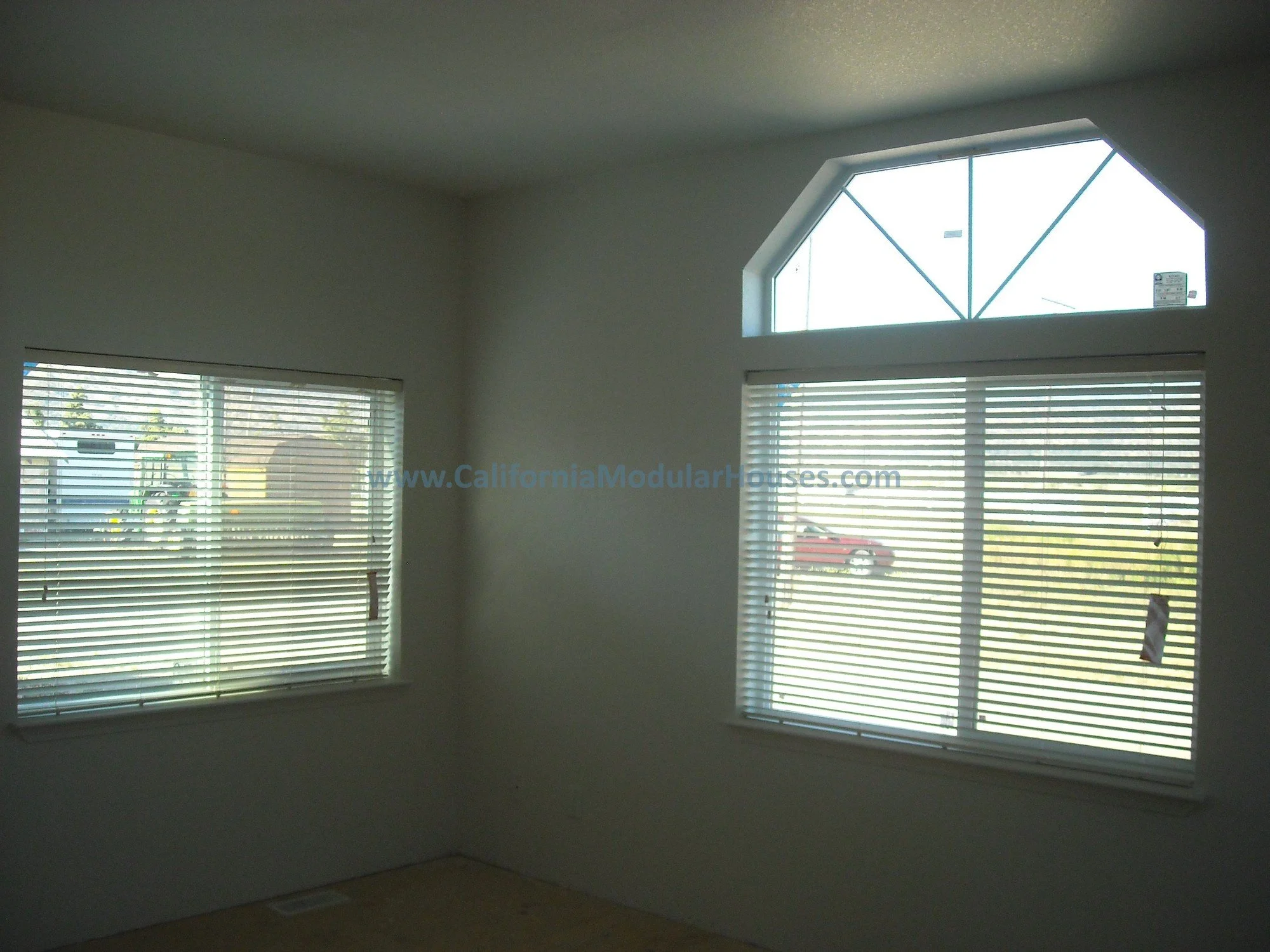 Interior of a room with three large windows, two rectangular with horizontal blinds and one semi-arched with a structural pattern, and a vent on the floor near the corner.  Look at all of the light coming into this home!  