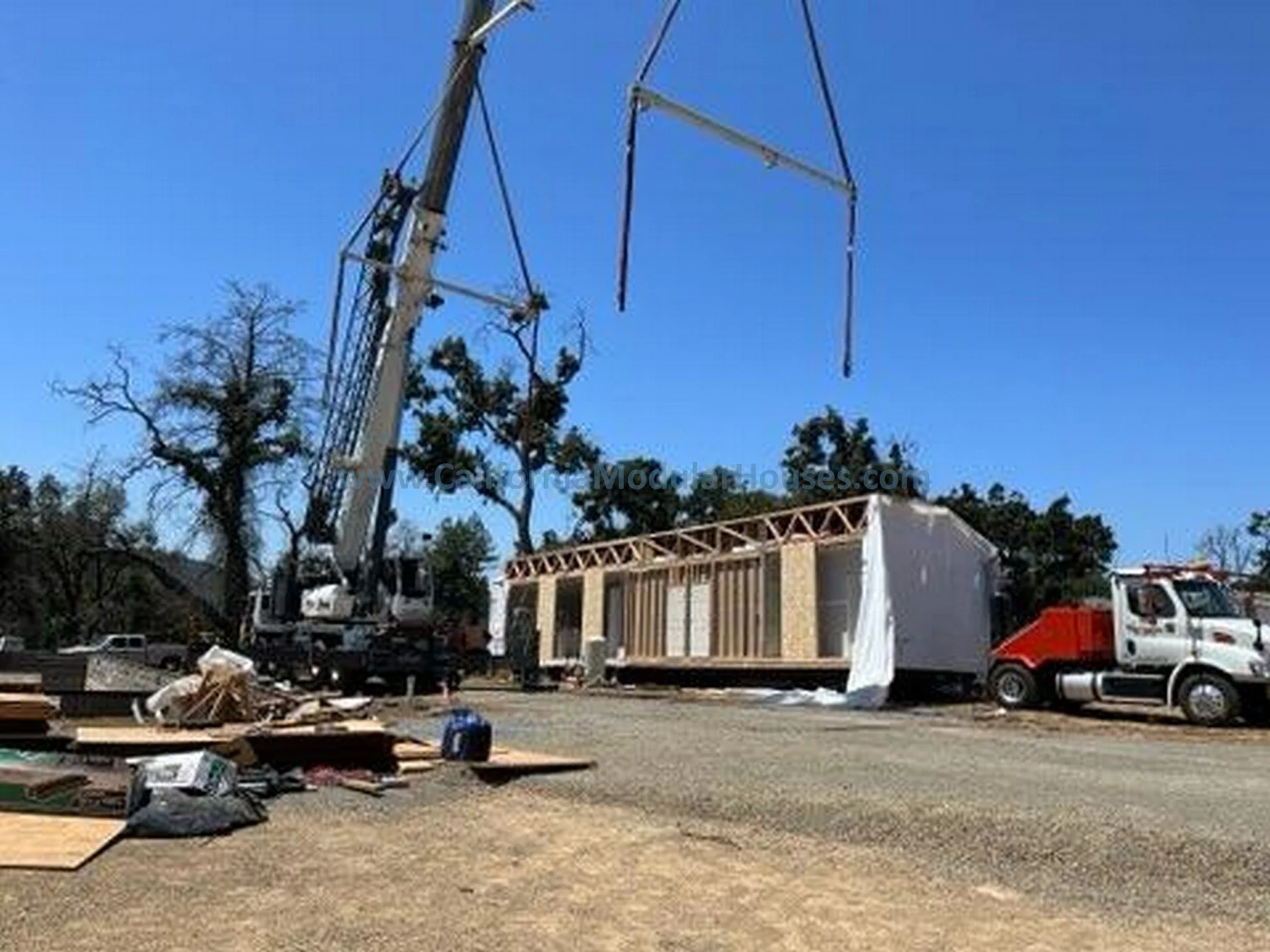 Construction site with a crane lifting building materials, a partially built house, and trucks parked nearby under a clear blue sky.
