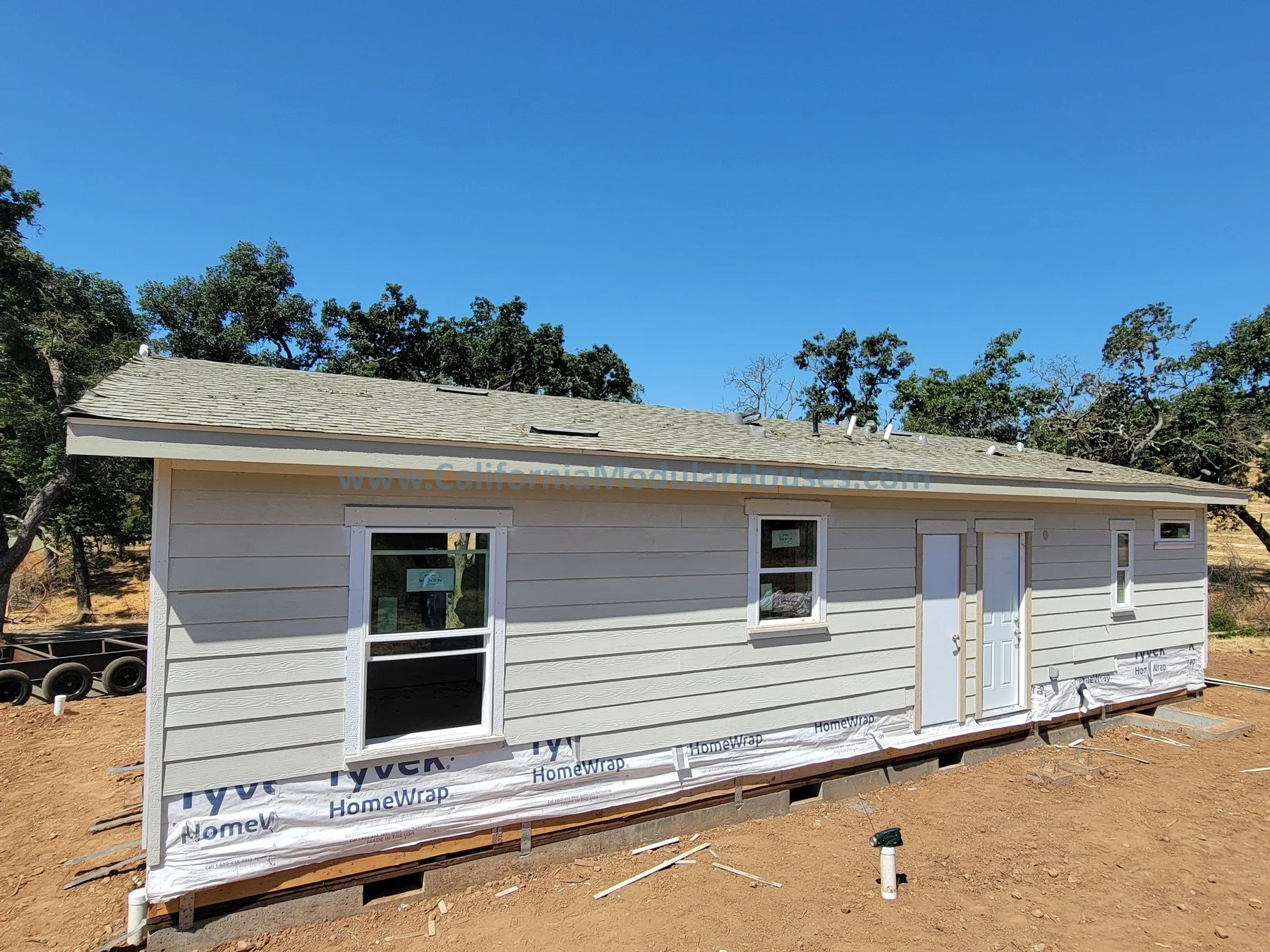 A small, recently constructed white house with siding, built on a raised foundation with support blocks. The house has several windows, a front door, and a side door, with a shingled roof and a clear blue sky above. The area around the house is bare 