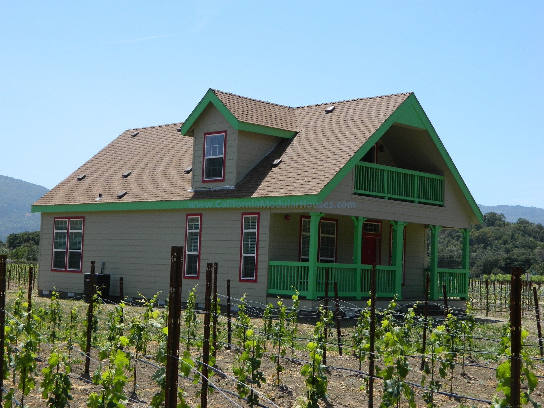 A two-story house with beige siding, green trim, and a gabled roof, located in a vineyard with rows of grapevines. The house has a front porch and a balcony, with rolling hills in the background.