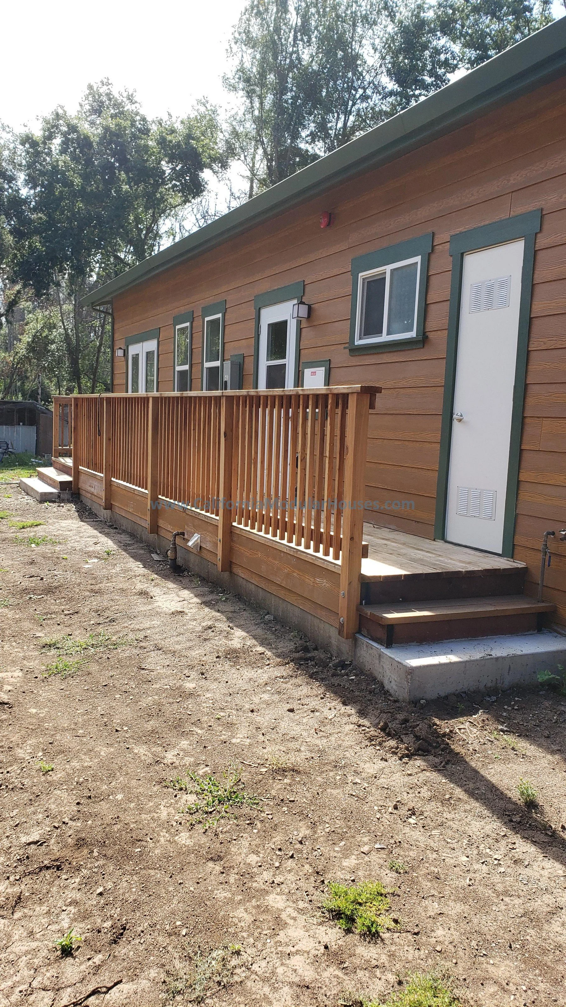A small wooden building with a metal roof, multiple windows, a white door, and a wooden deck with stairs and a railing, set on a dirt lot with some small patches of grass. Prefab Modular Accessory Dwelling Unit