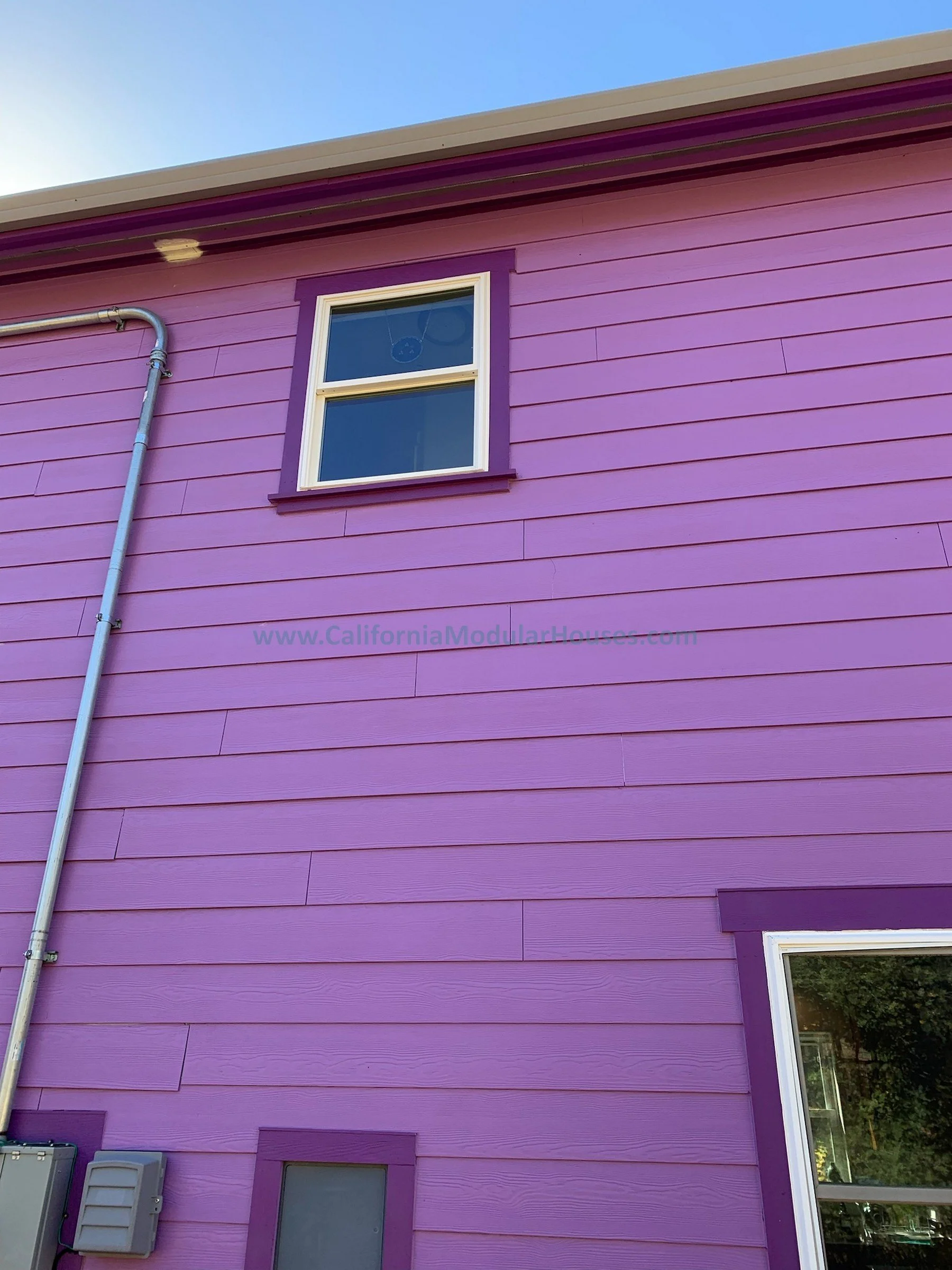 Close-up view of a purple house's exterior wall with a double-pane window and purple trim, a gray gutter and downspout, and utility boxes at the bottom.  Modular Home CA, Modular Homes, Pre-Fabricated Homes,